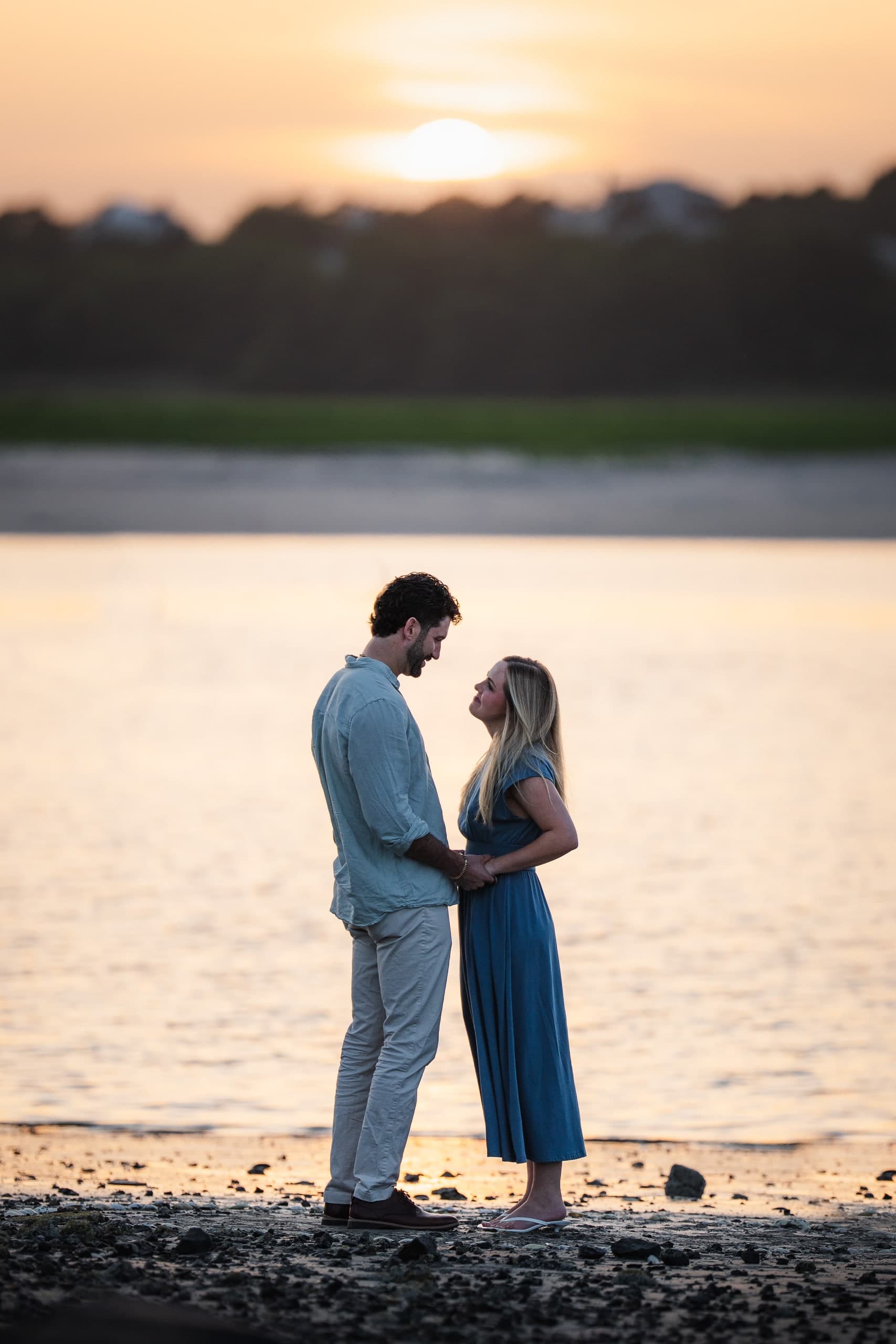 A couple facing each other and holding hands on the shoreline, sun setting behind them.
