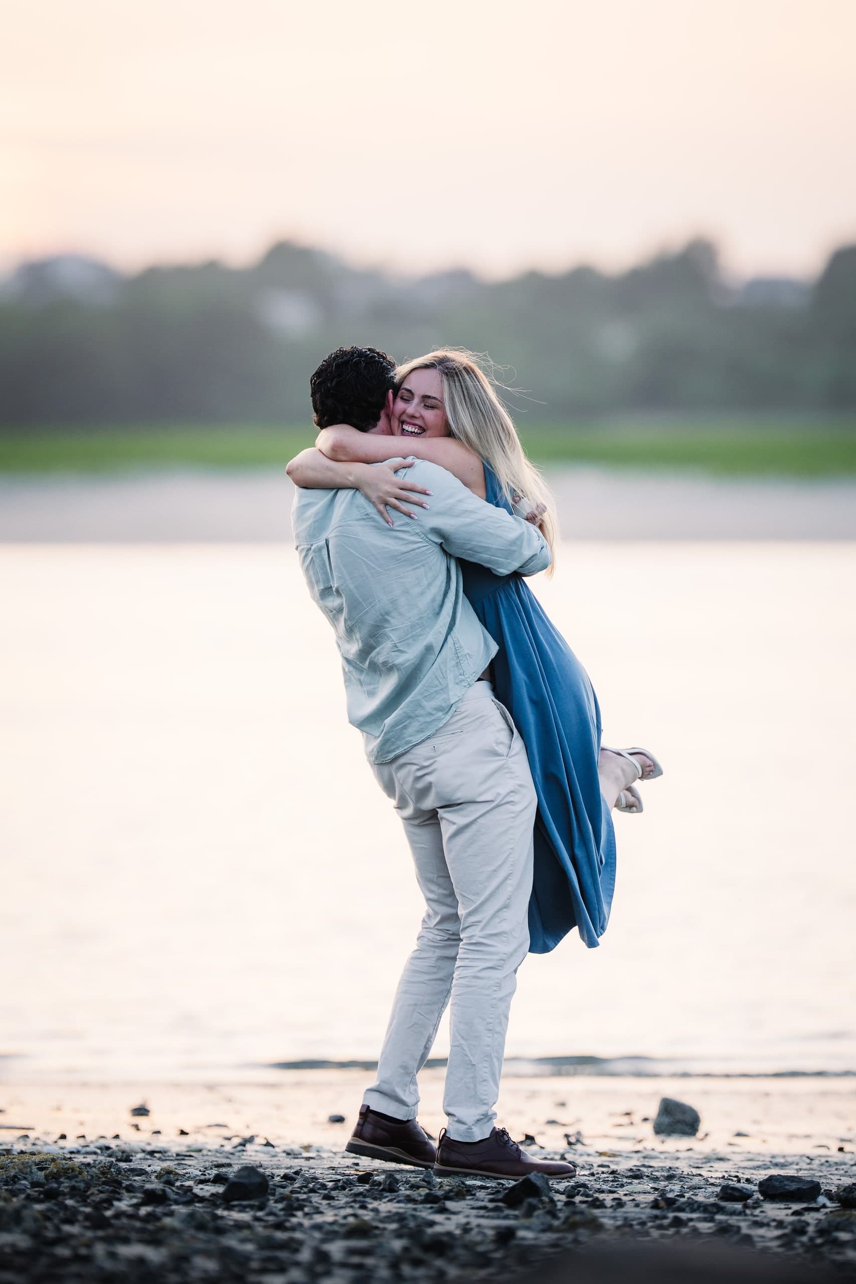 A man lifting his newly engaged partner off the ground, both laughing, at dusk.