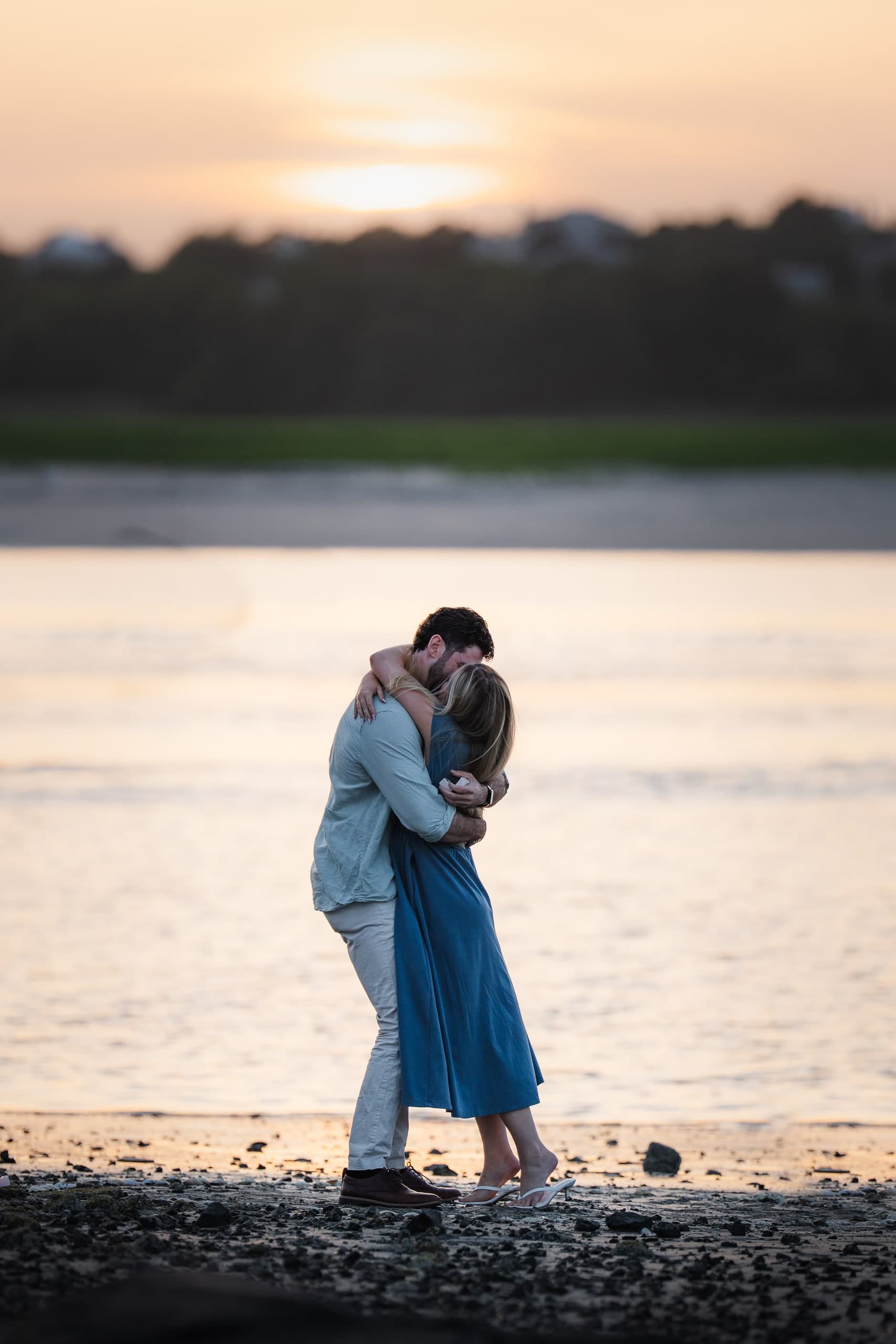 A couple kissing on the shoreline at sunset, standing close, water and sky behind them.