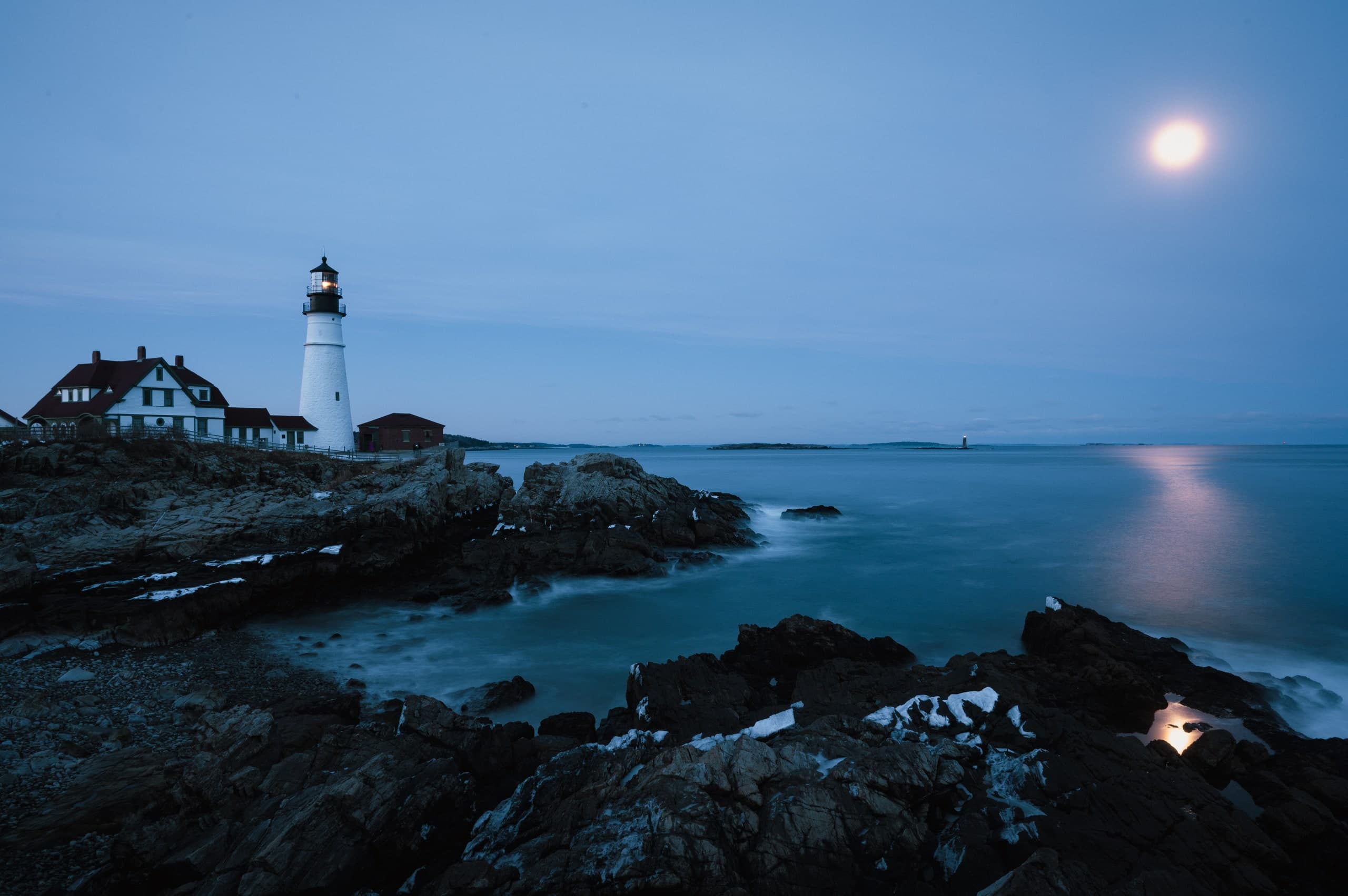 A lighthouse on a rocky New England point at dusk, a full moon rising over the water.
