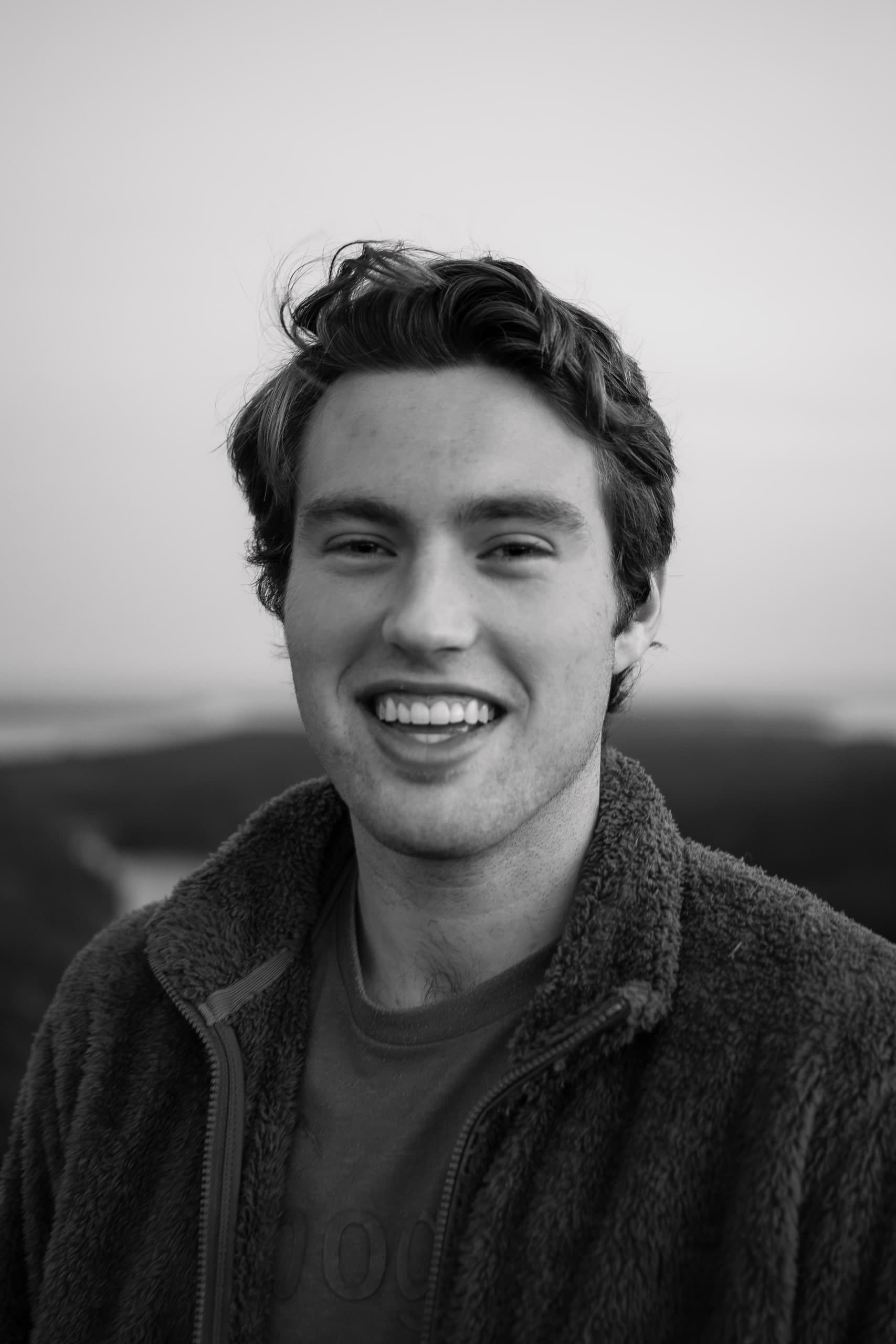 Black and white portrait of Oliver Spivey, smiling, outdoors with the coast in the background.