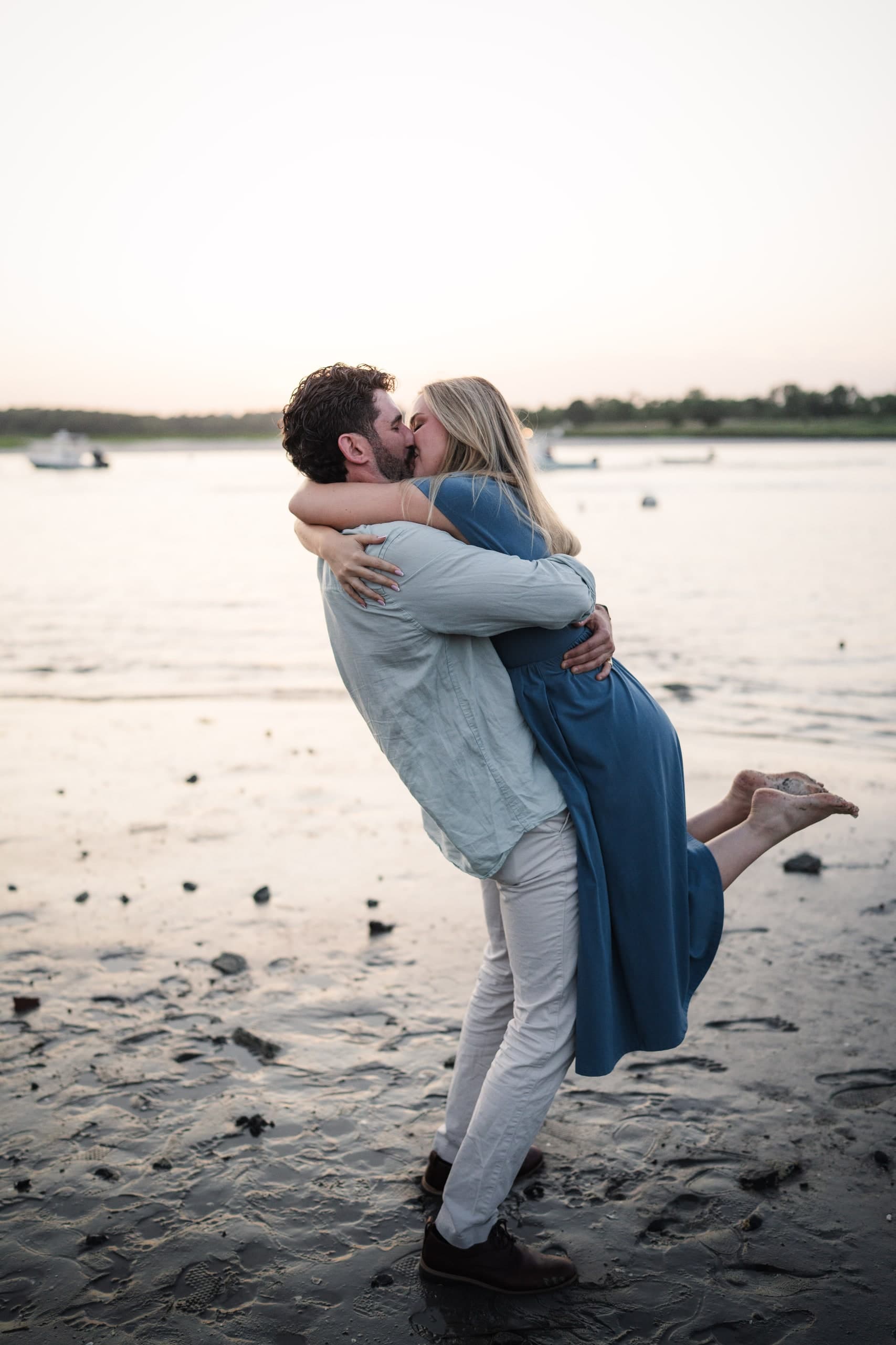 A man lifting his newly engaged partner into a kiss on the shoreline at sunset.