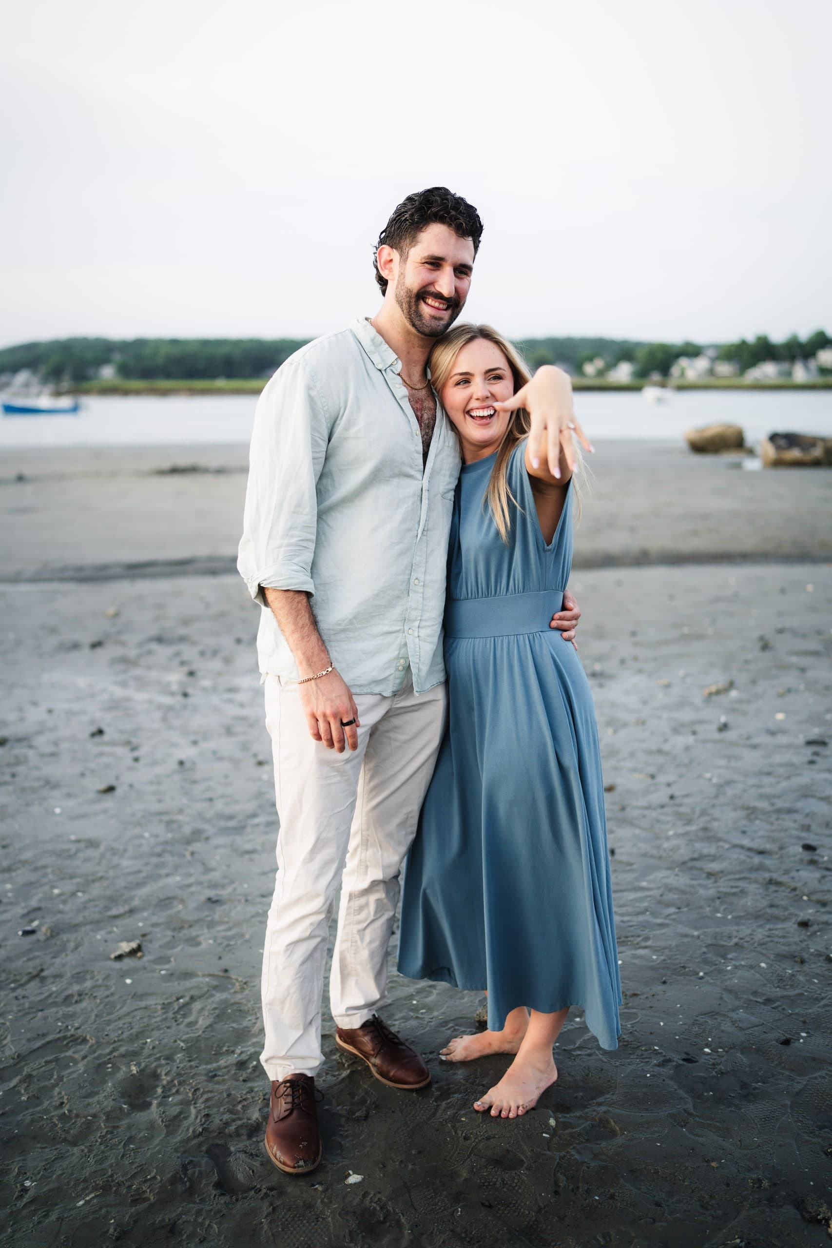 A newly engaged couple laughing together on the beach, his hand raised in celebration.