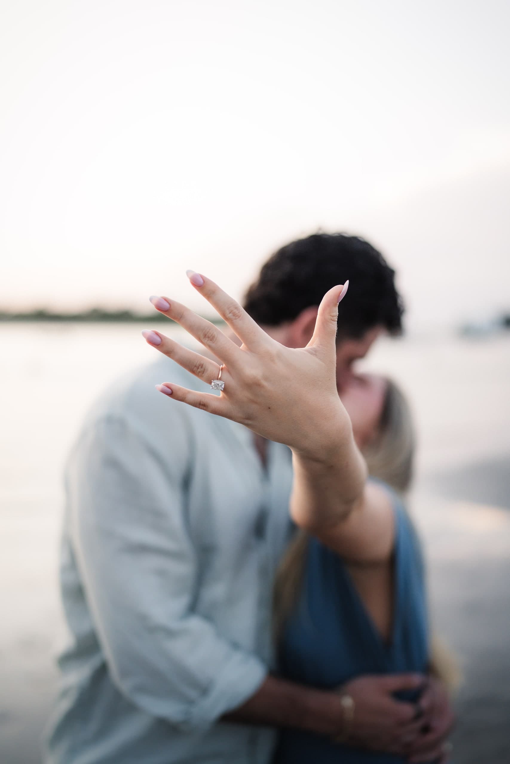A newly engaged couple embracing on the beach at dusk, her ring hand raised.