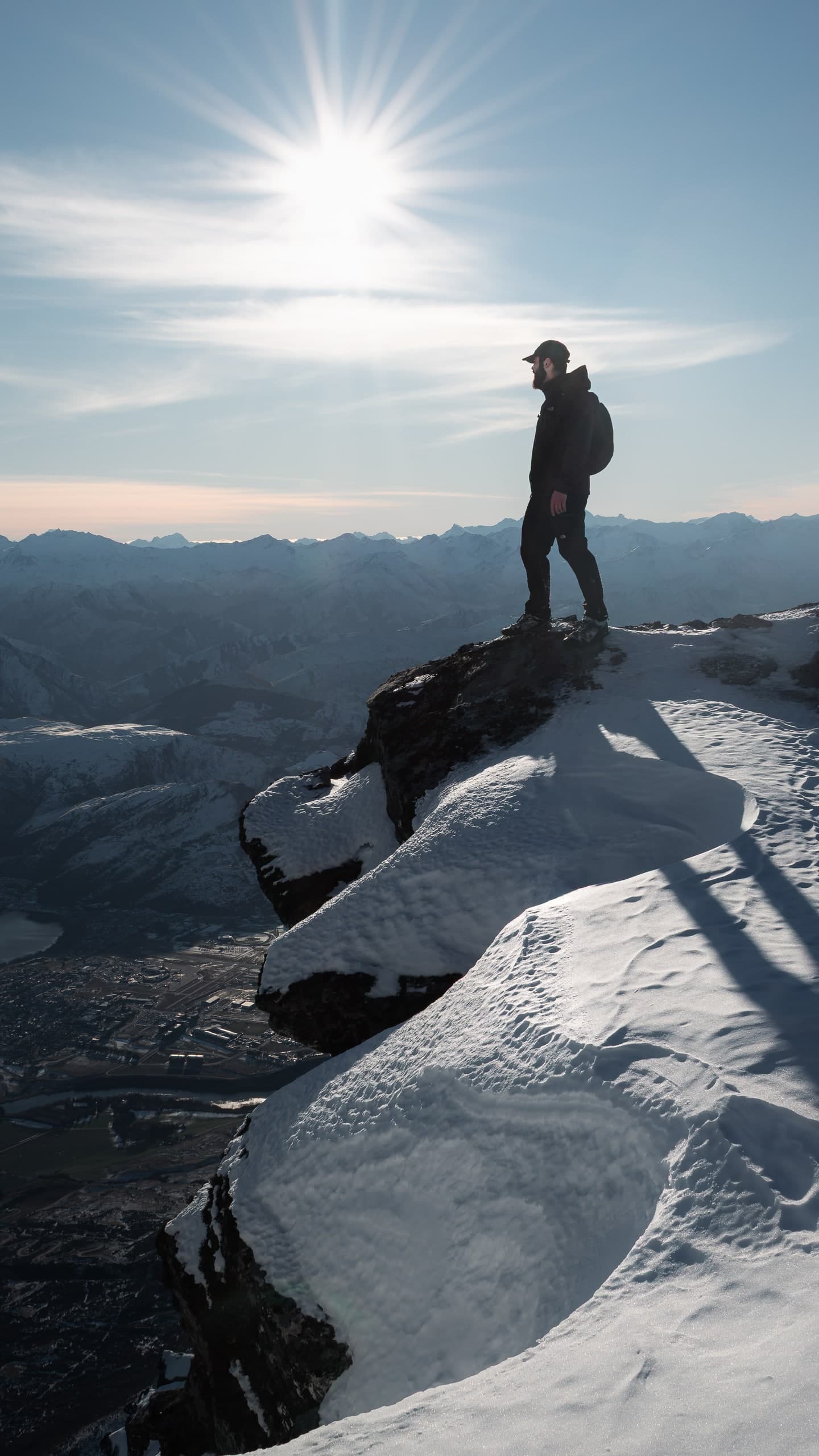 A hiker standing on a snowy mountain summit, sun low over distant peaks.