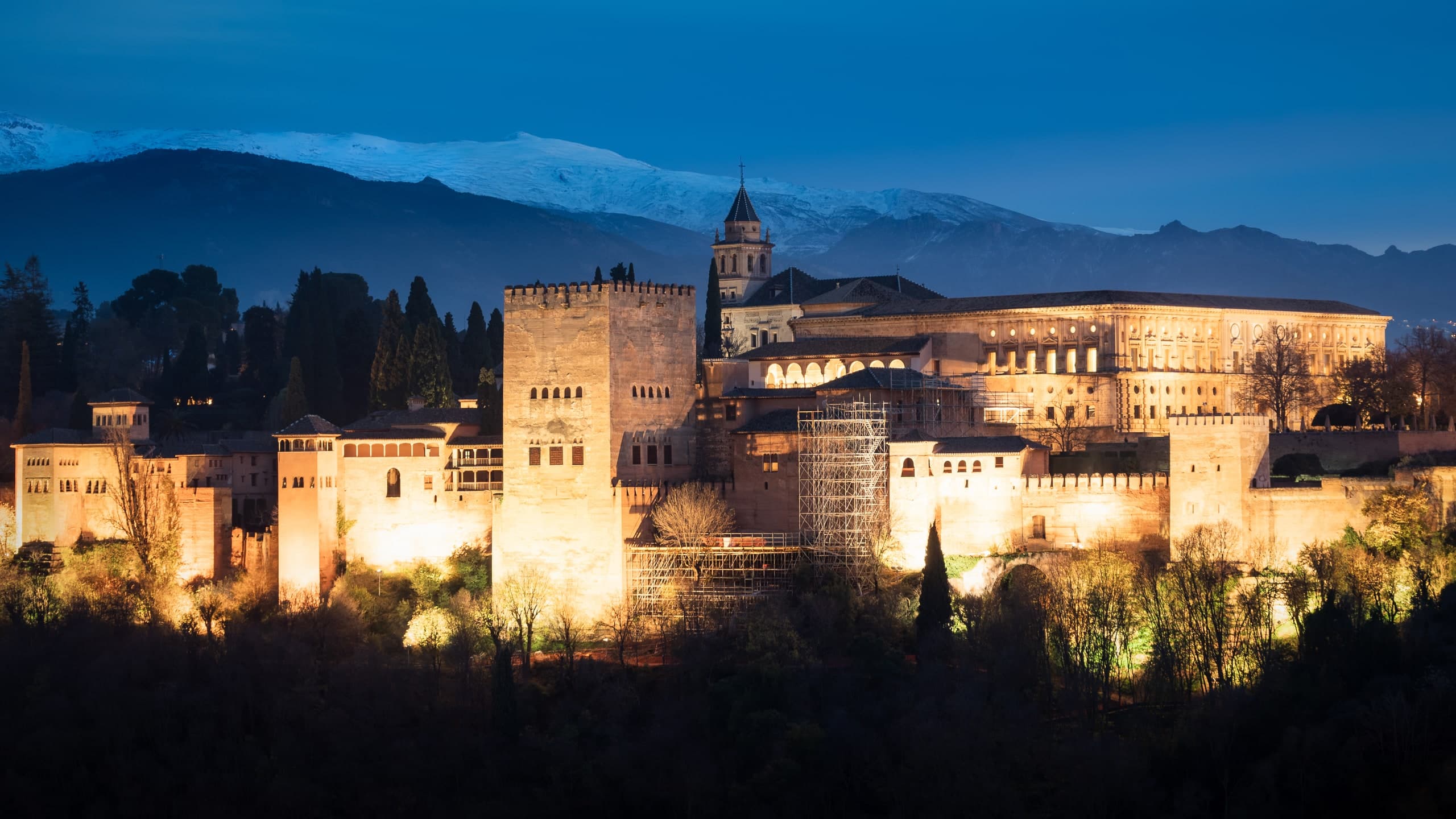 The Alhambra lit at night, snow-capped mountains behind it.
