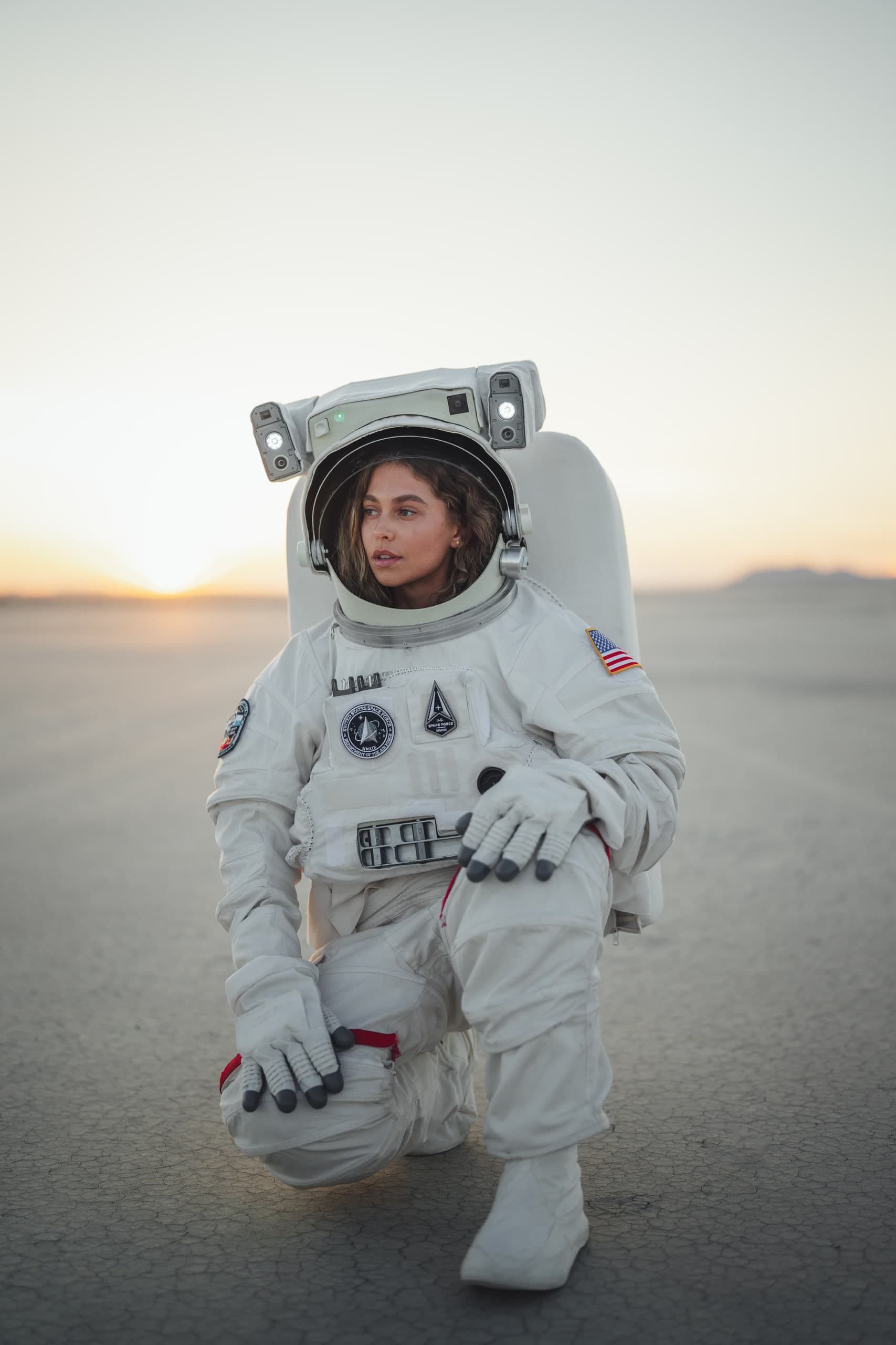 A woman in a full astronaut suit kneeling on a dry lakebed at sunset.