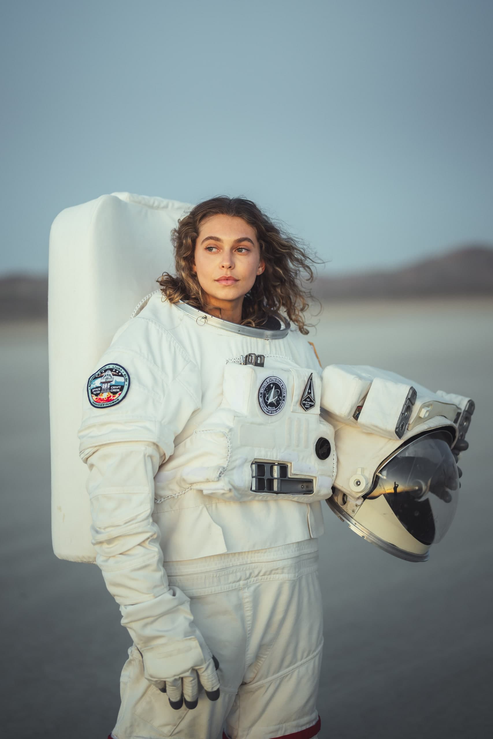 A woman in a full astronaut suit standing in profile, holding her helmet, dusk light on a dry lakebed.