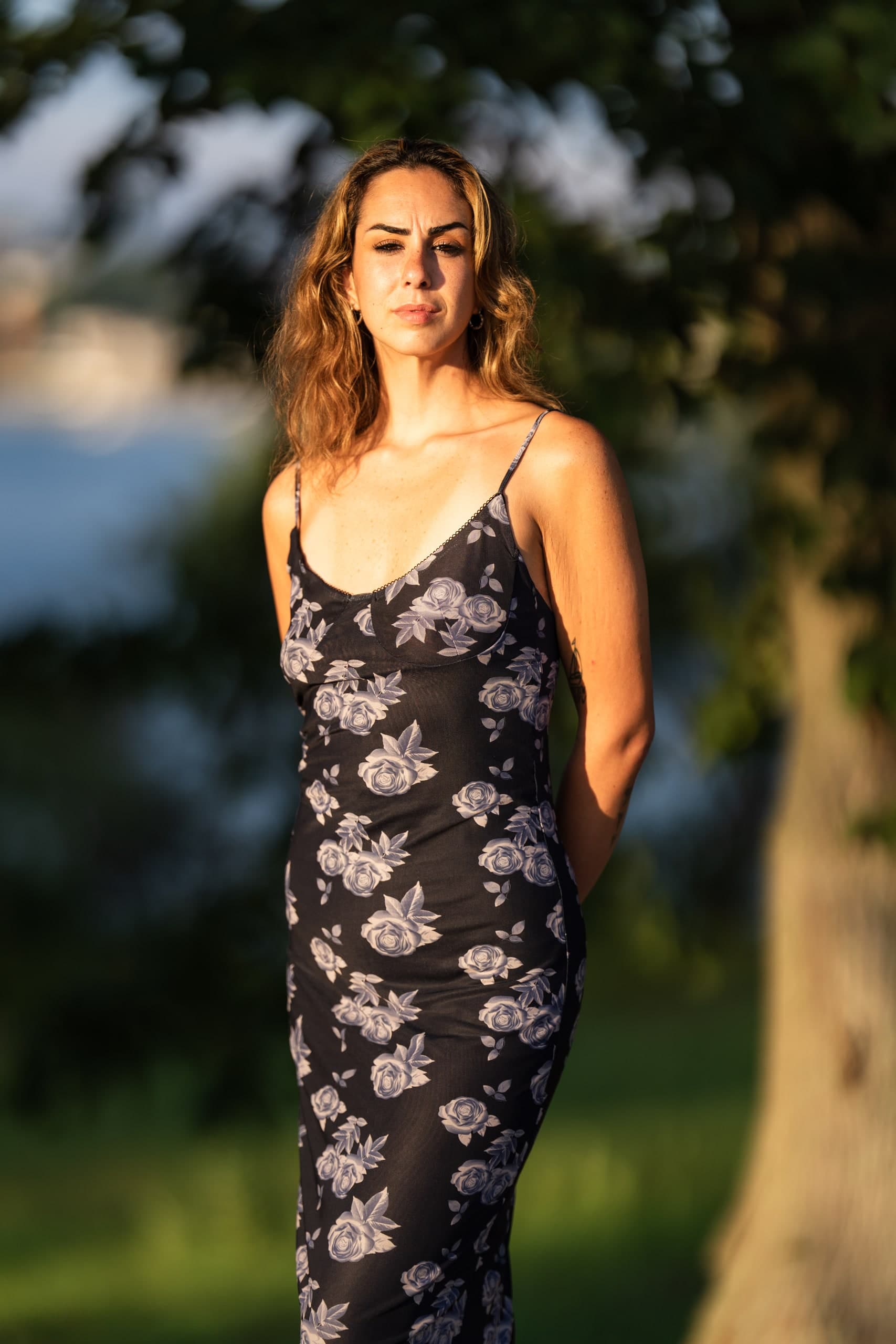 A woman in a dark blue floral dress standing in a field, looking directly into the camera with a focused expression.