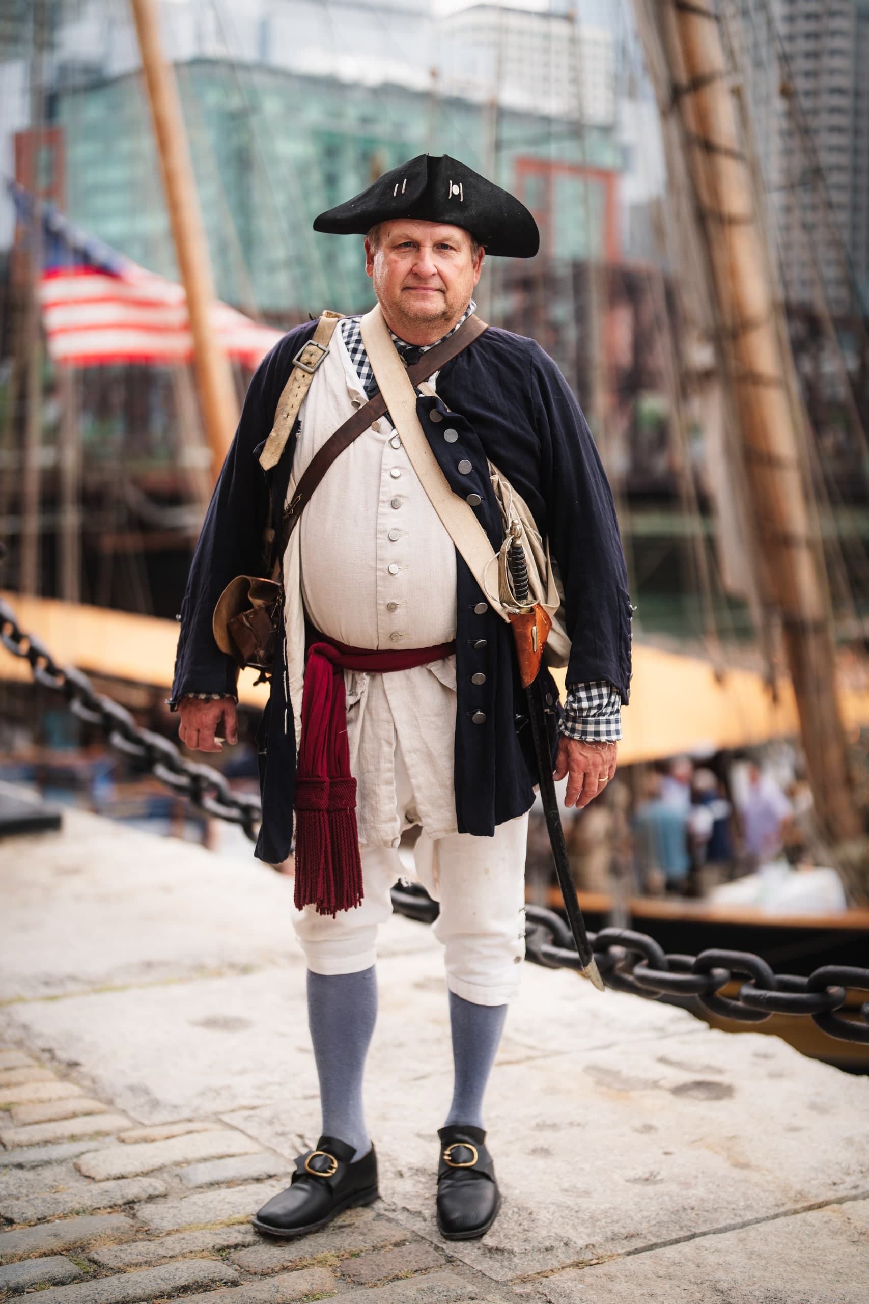 A man in full colonial military reenactment costume standing on a tall ship dock, city skyline behind him.