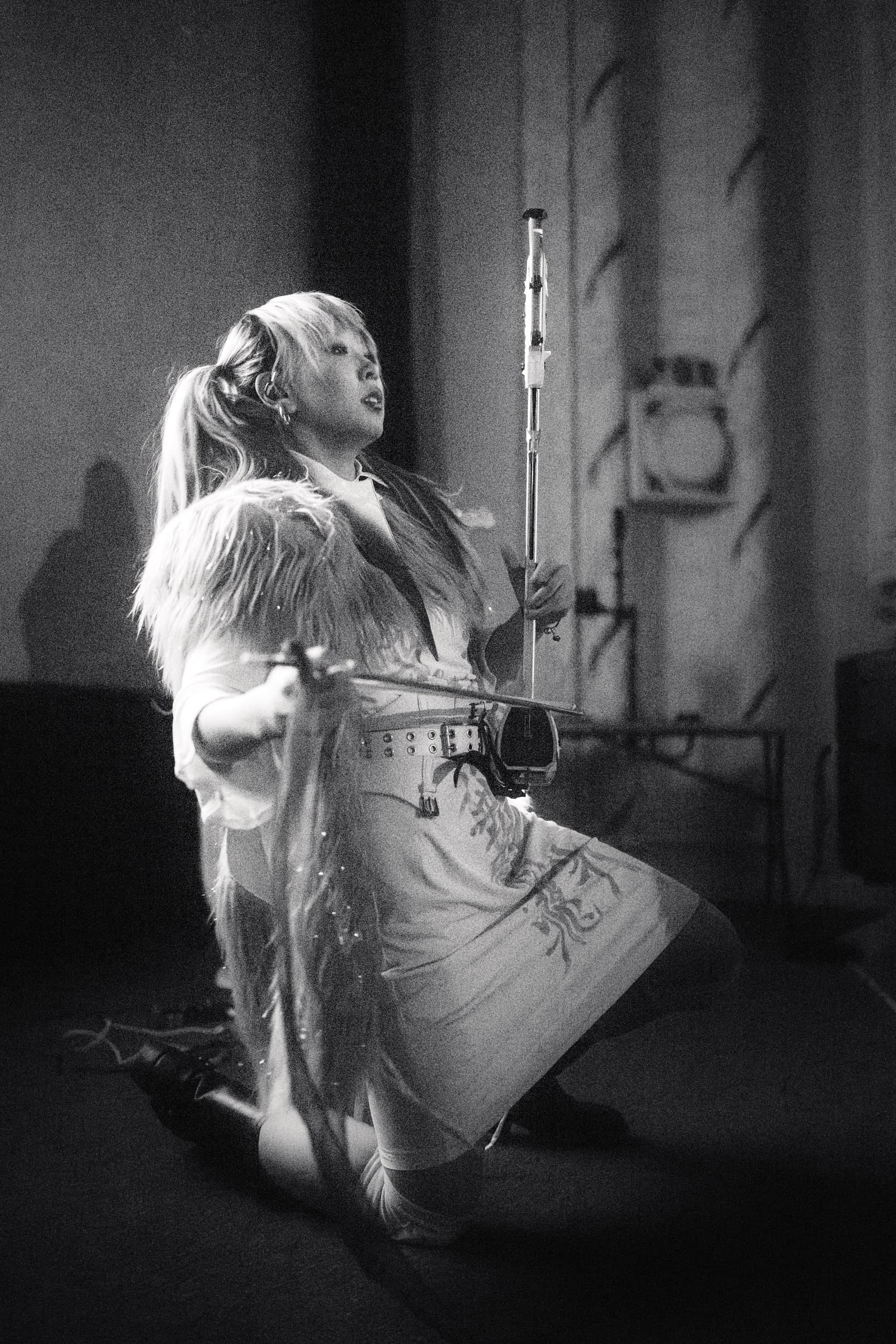 A black-and-white photo of a performer in a feathered costume on stage, spotlit against a stairwell.