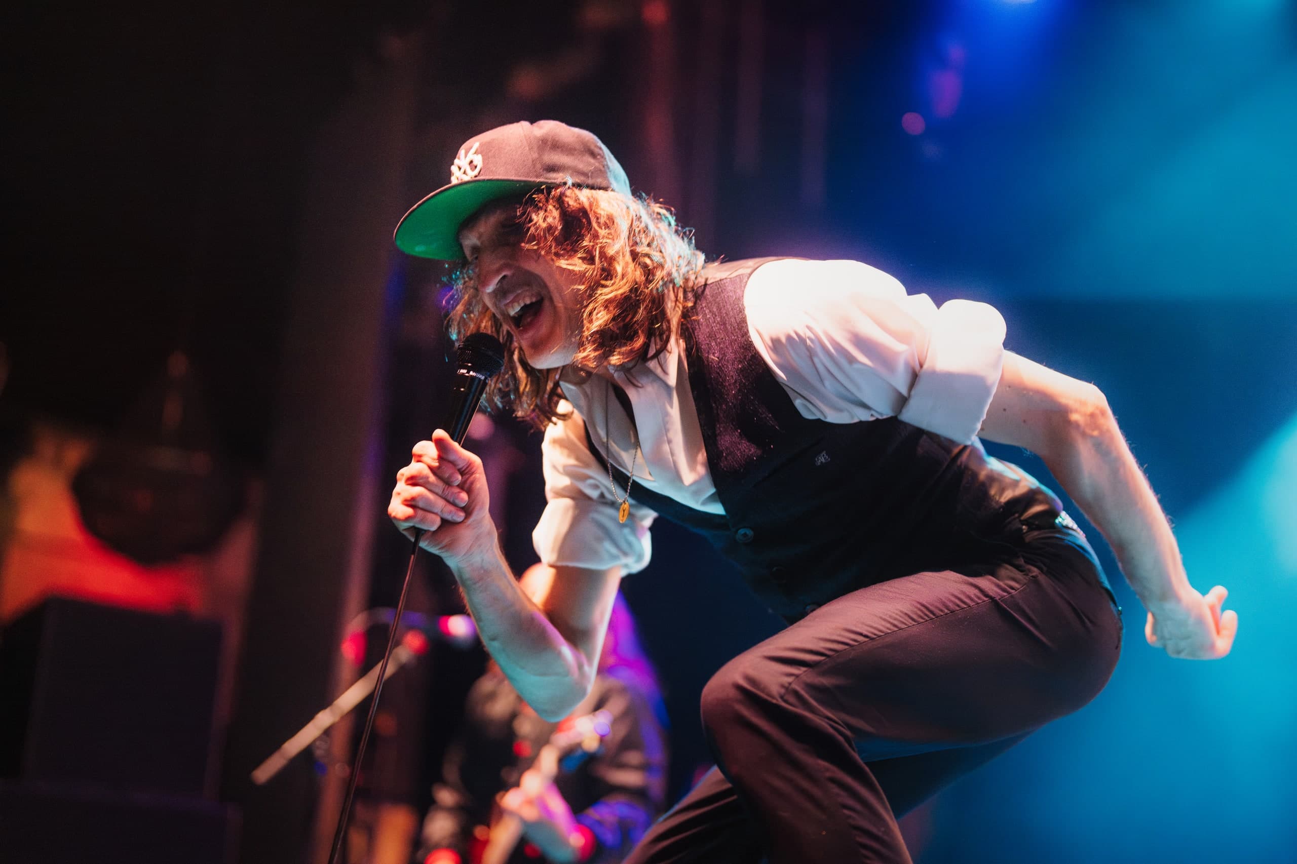 A singer in a cap leaping mid-performance under blue stage lights.