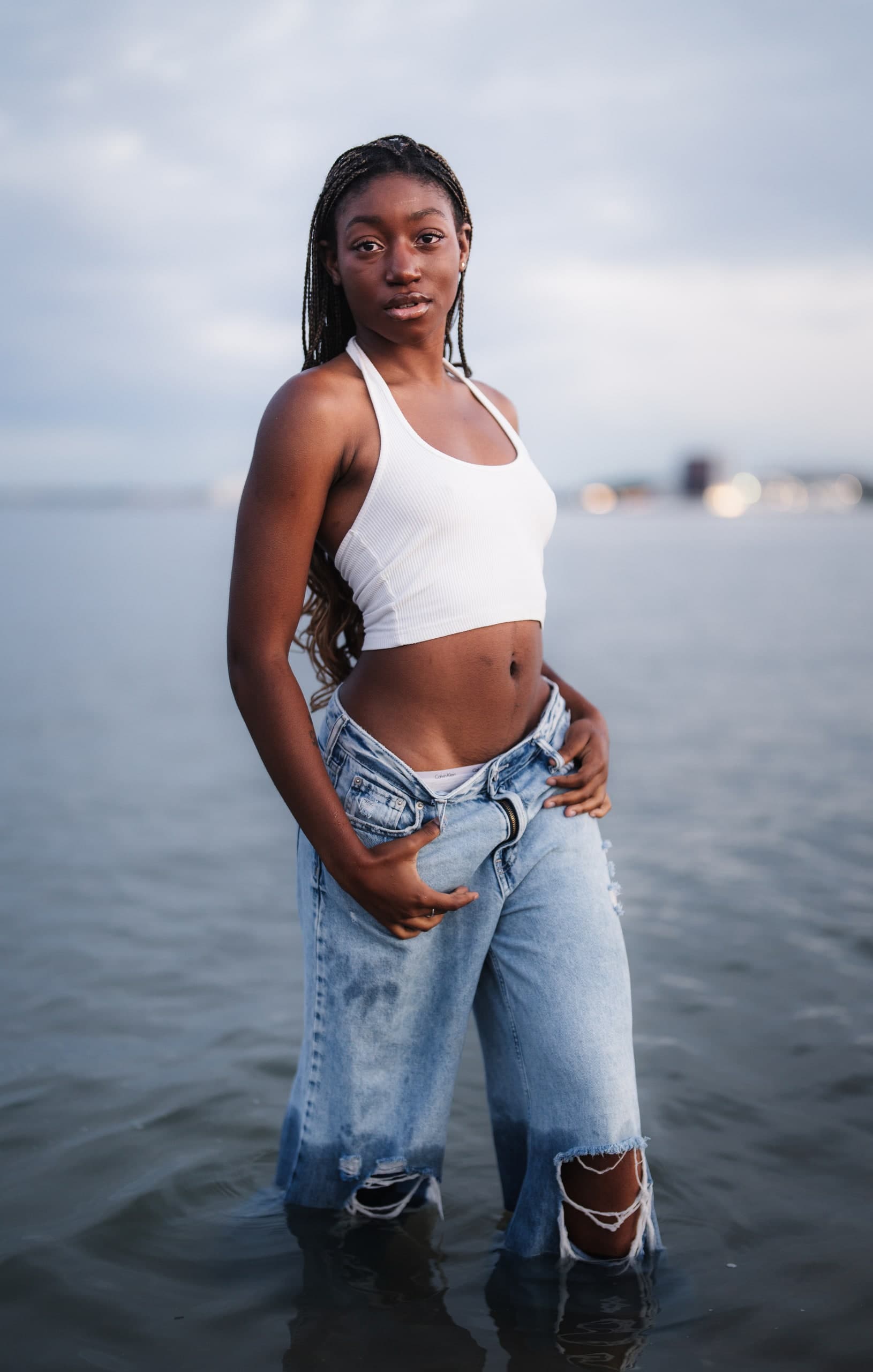 A woman in denim and a white top standing knee-deep in the water, city lights behind her.