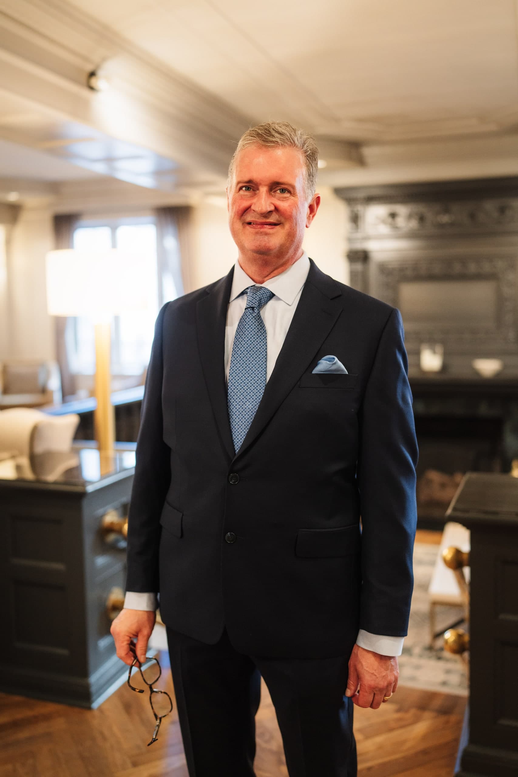 A man in a navy suit with a blue tie and blue pocket square, smiling indoors.