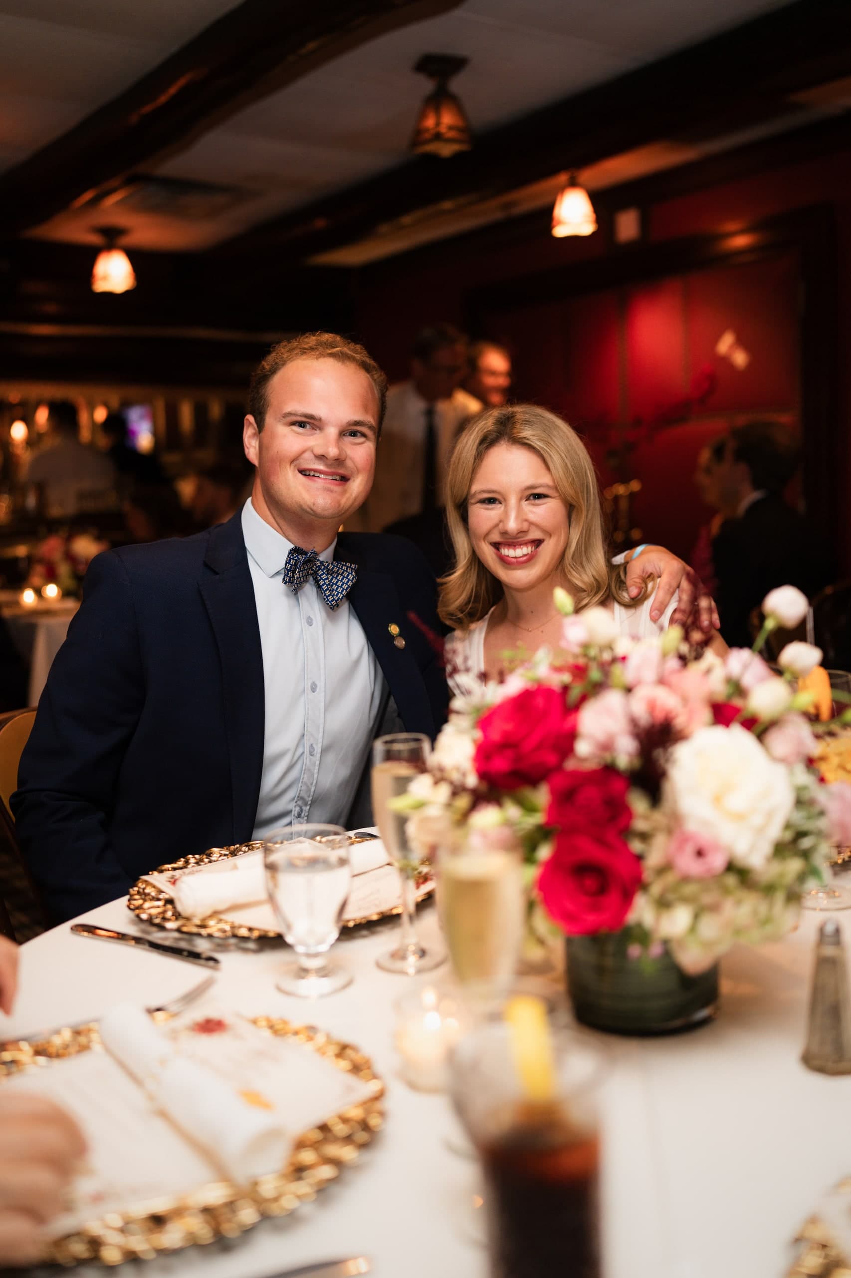 A couple smiling with an arm around each other at their table, a bouquet of red and pink roses beside them.