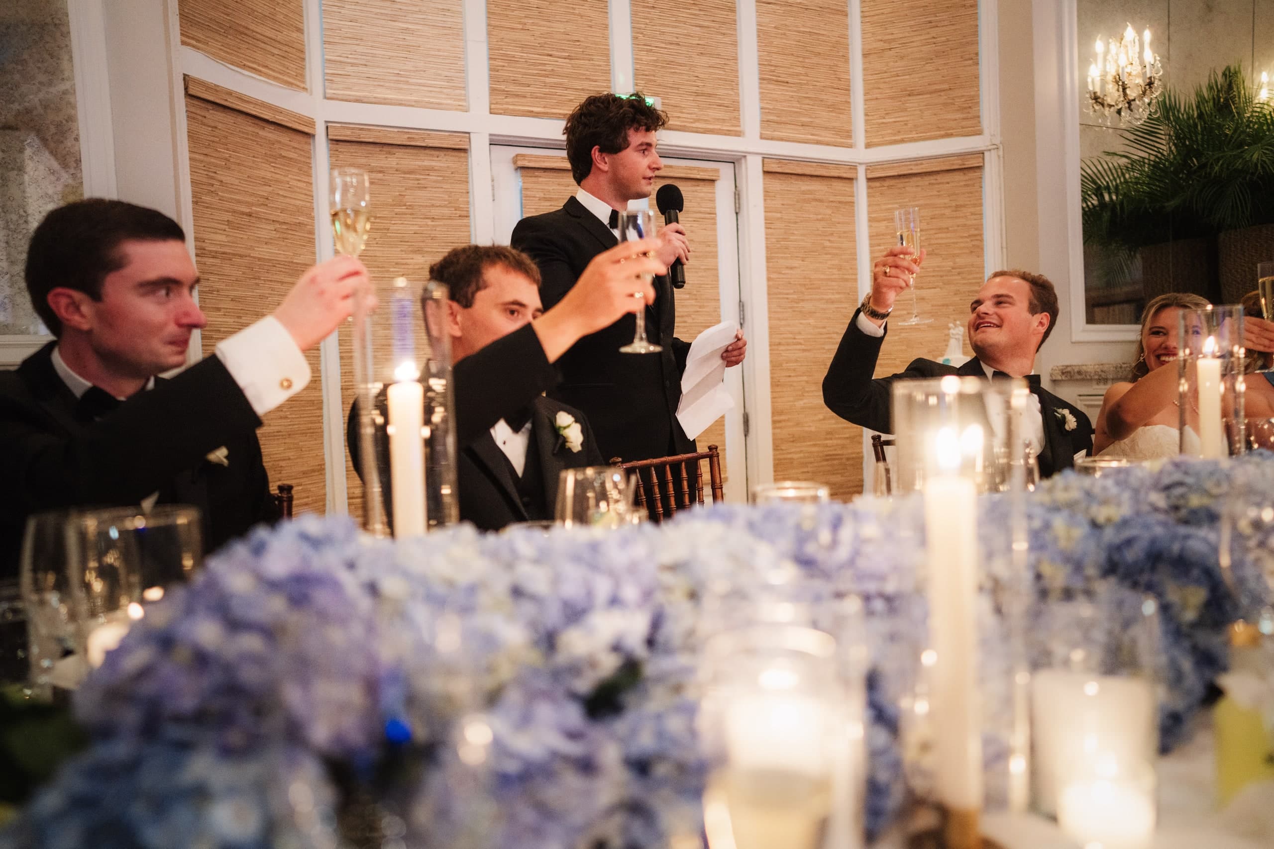 A groomsman giving a toast, glasses raised around the table, candlelight and blue hydrangeas in the foreground.