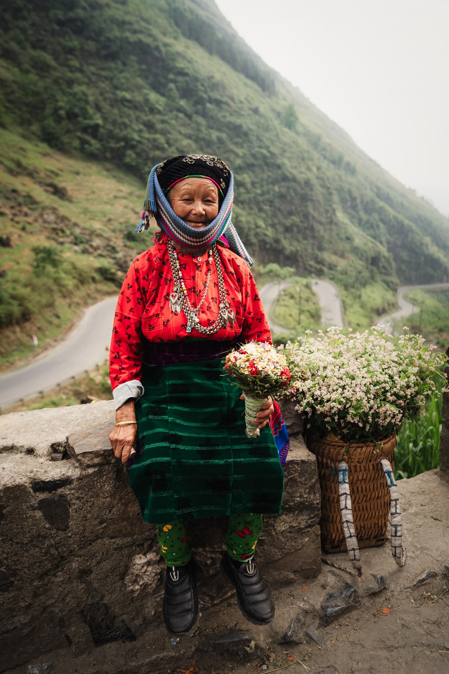 A woman in traditional dress holding flowers, standing on a mountain road.