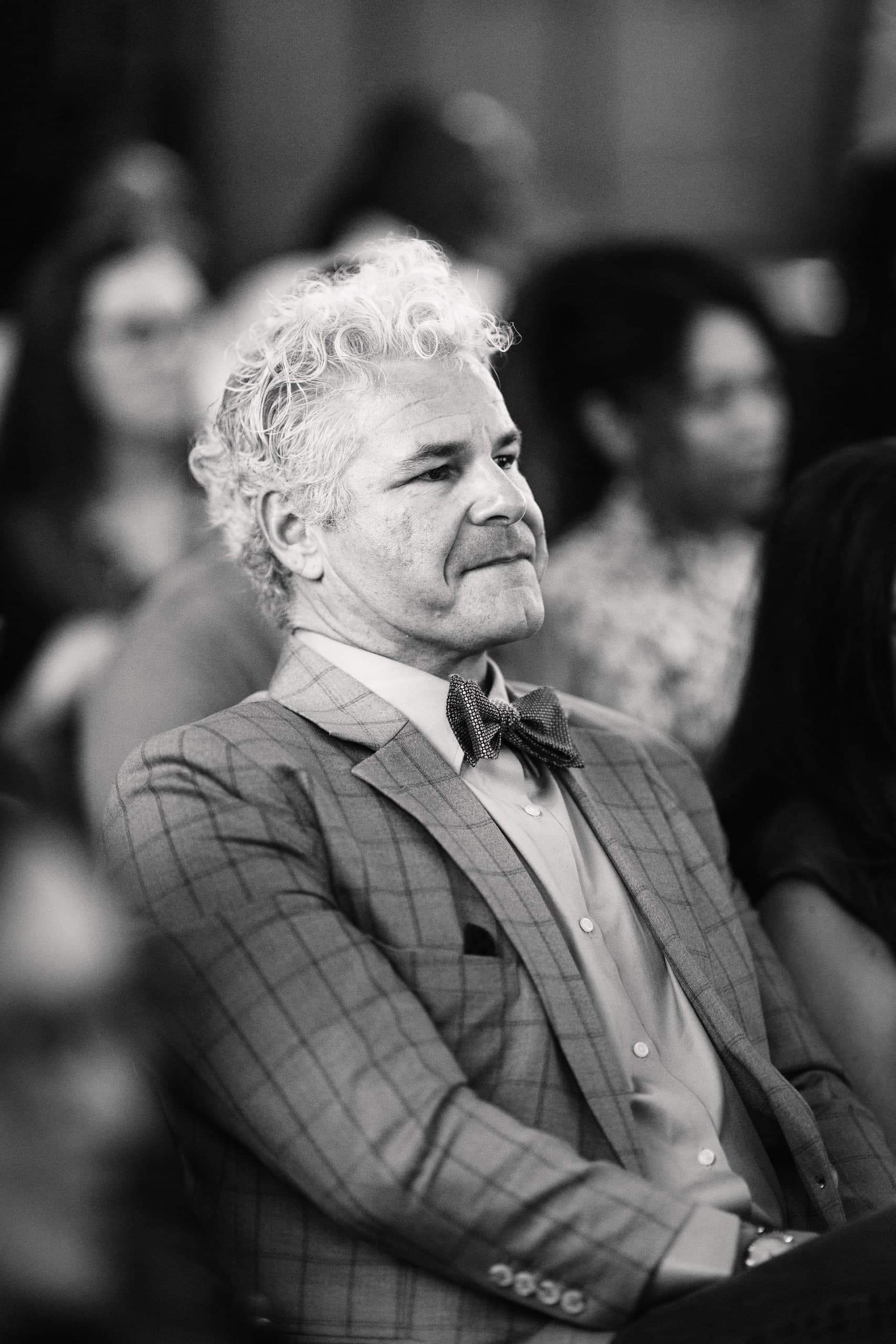 A black-and-white portrait of a man in a bow tie and plaid suit, smiling from a formal audience.