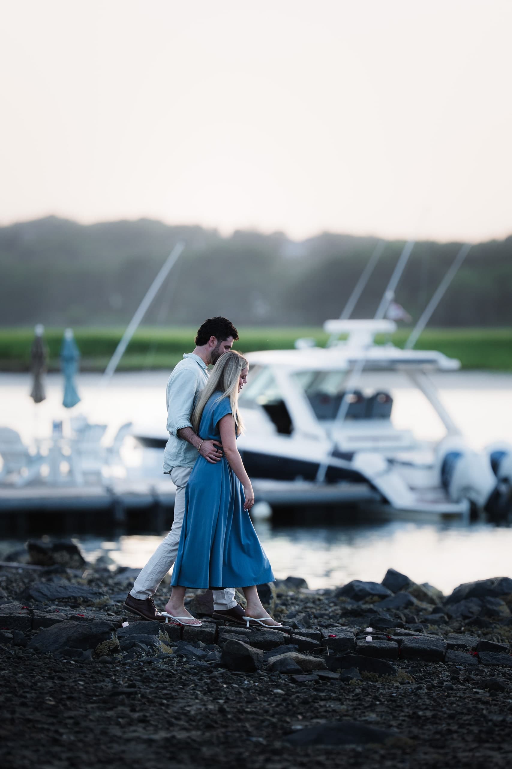 A couple walking along a dock at dusk, boats moored beside them, marsh grass in the distance.