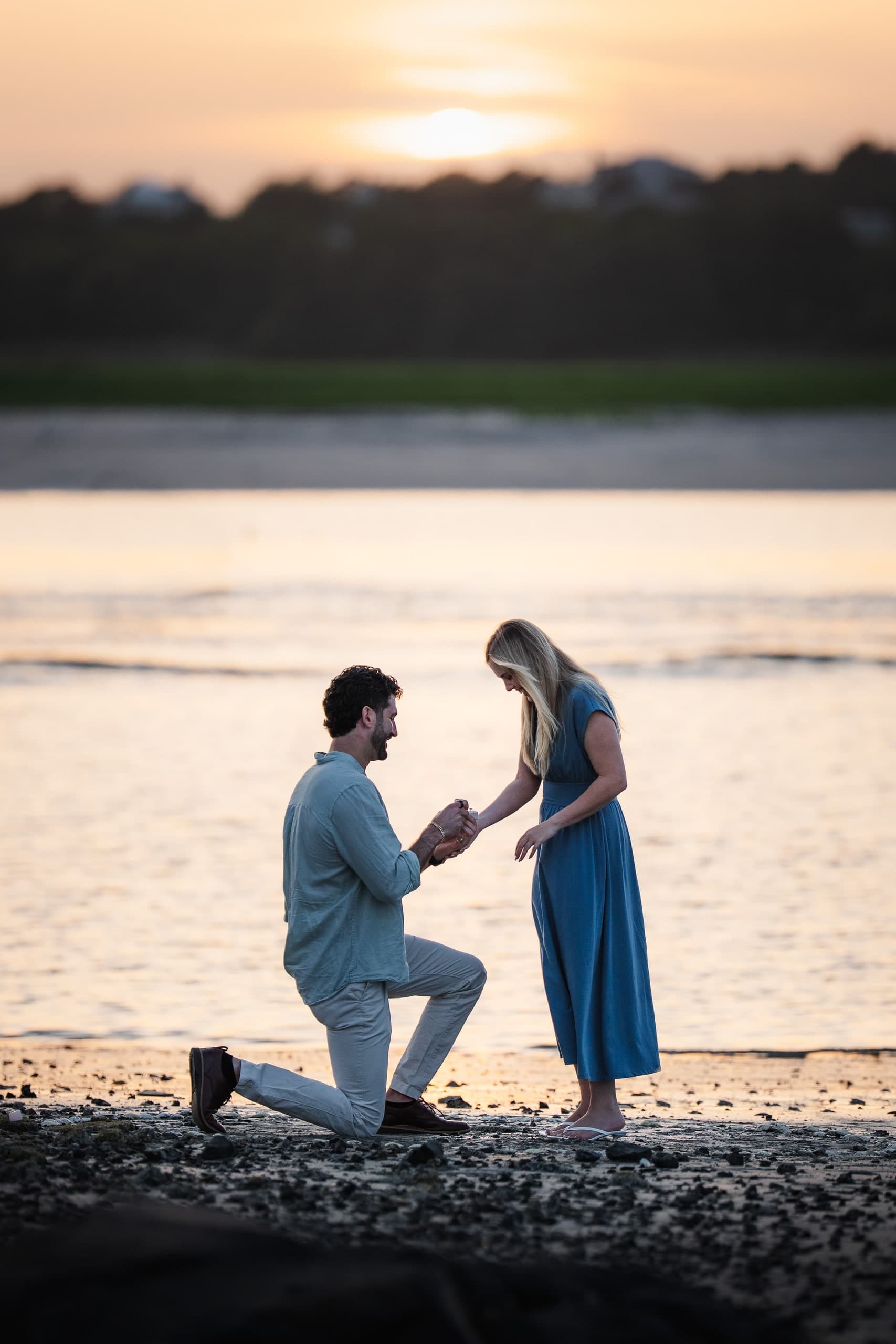A man kneeling on the beach, proposing, as the woman looks down at the ring.