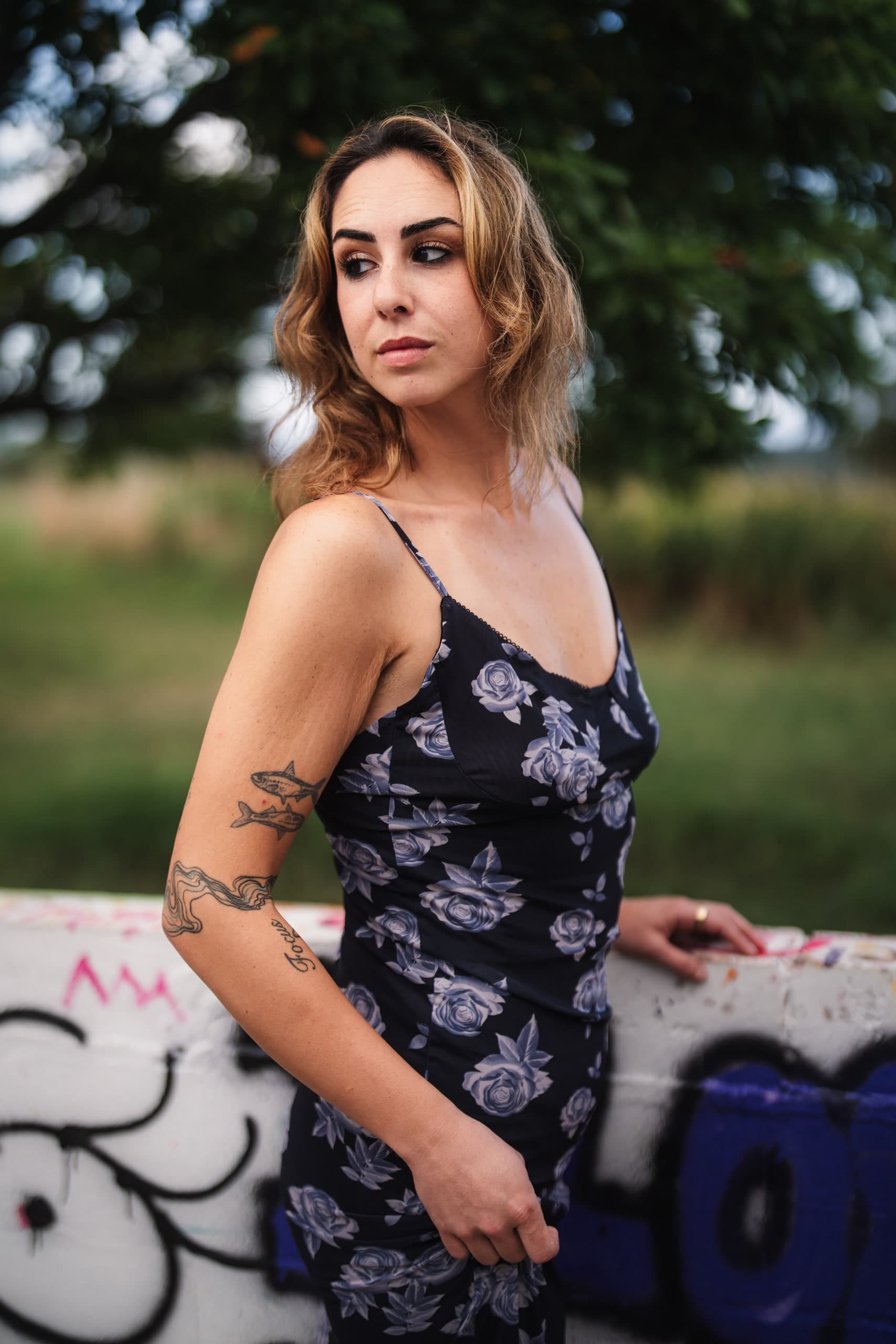 A woman in a dark floral dress looking back over her shoulder, leaning against a graffiti-covered wall.