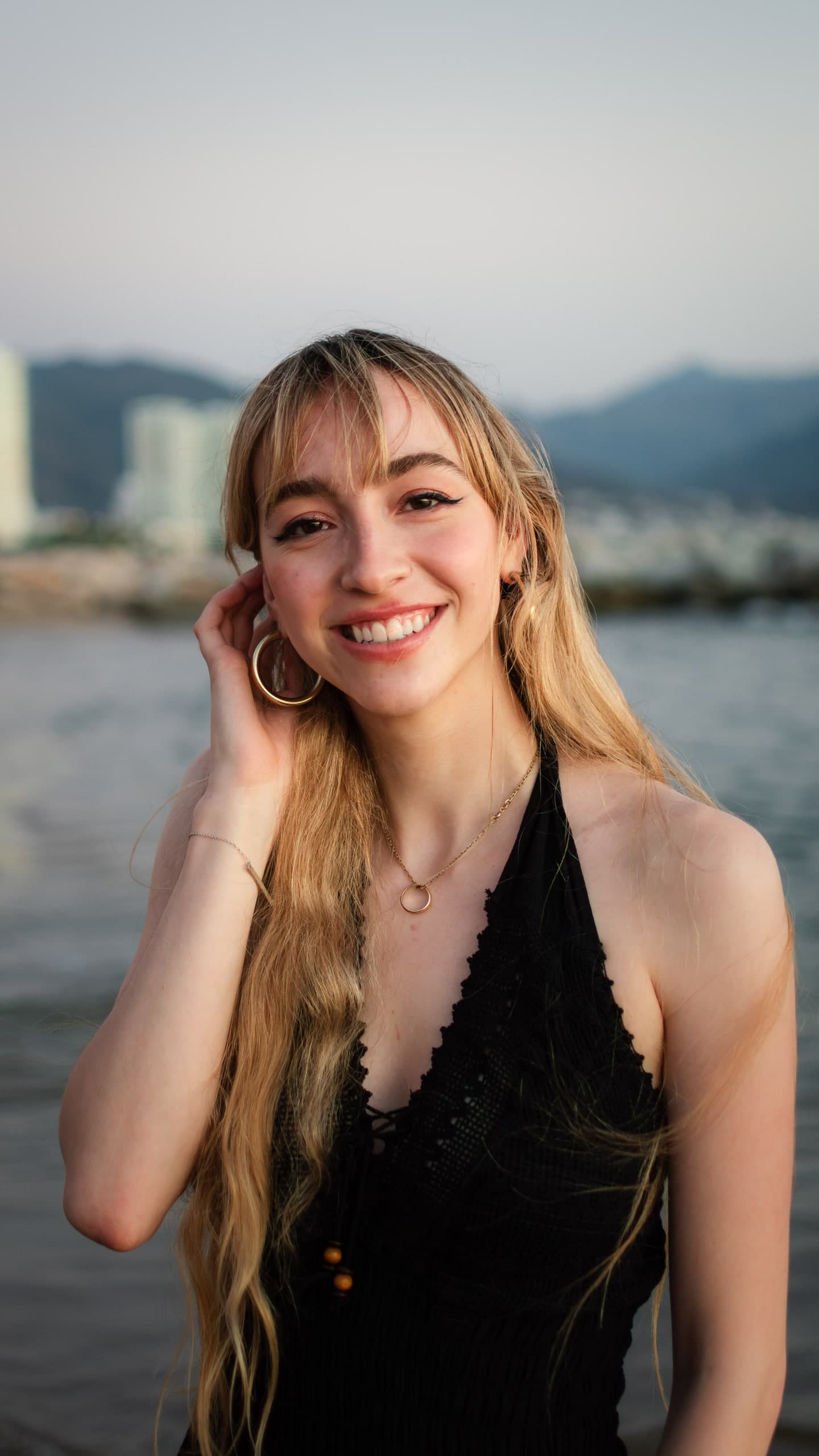 A blonde woman smiling, touching her hair, a city skyline and water behind her.