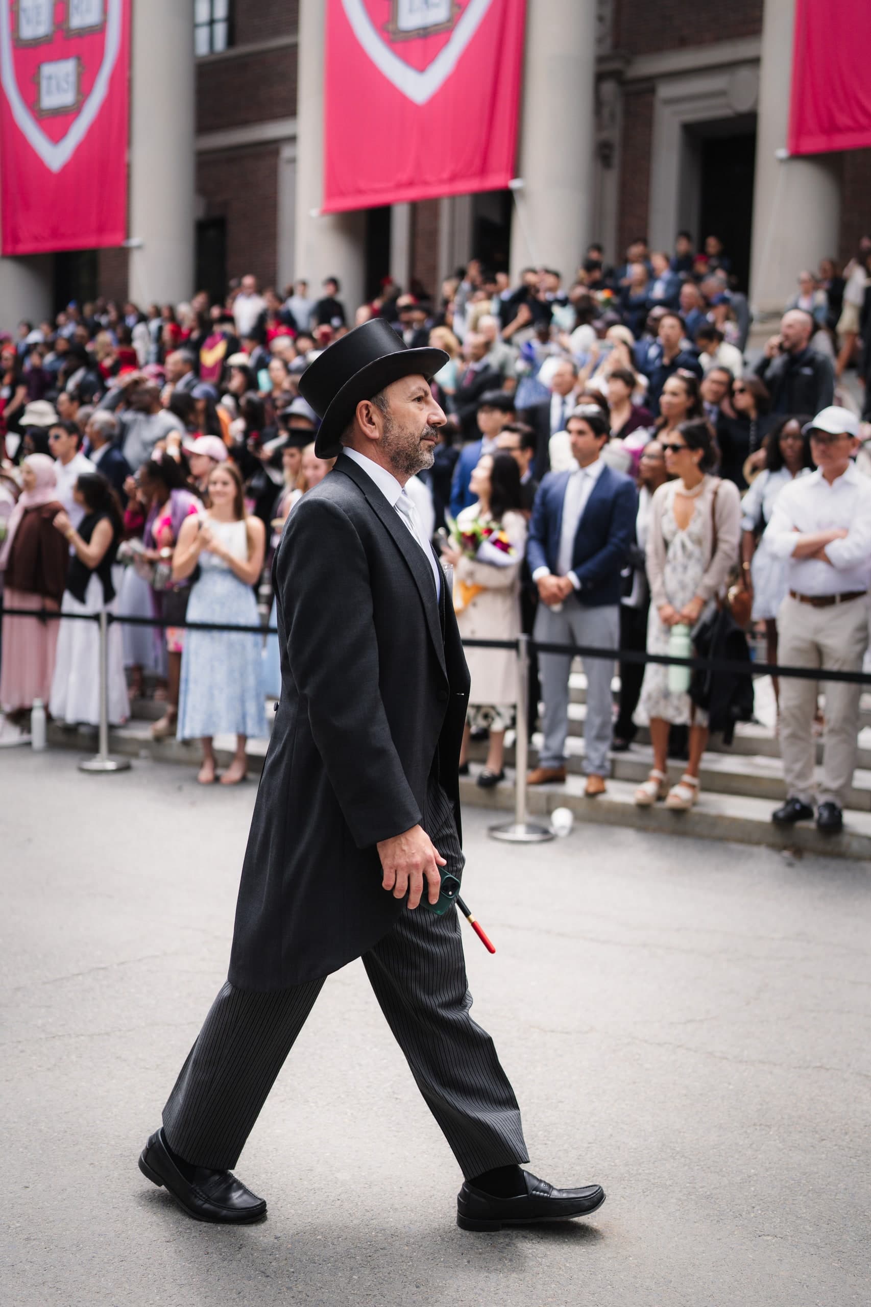 A man in a top hat walking in a Harvard commencement procession, red banners behind him.
