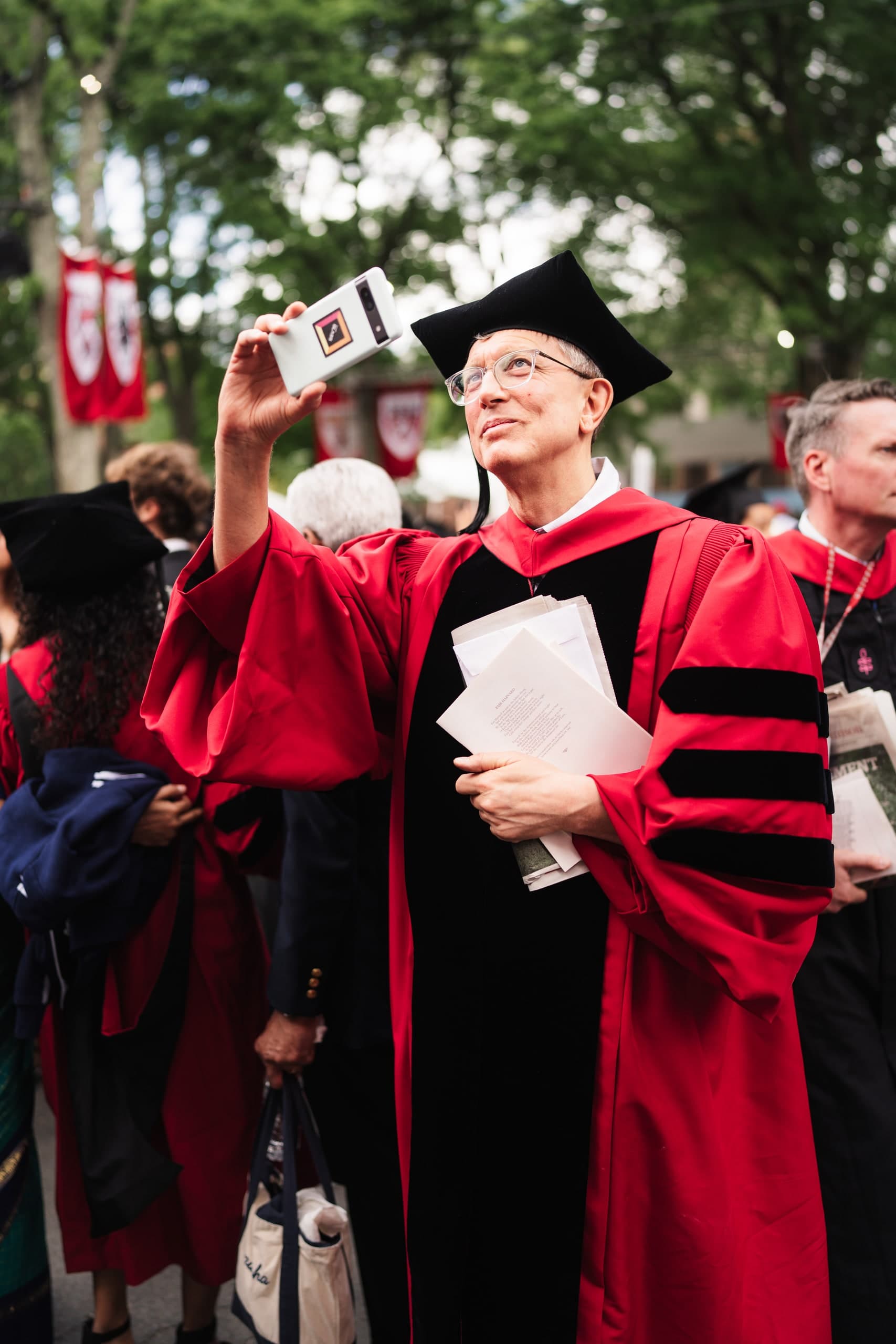 A professor in red doctoral robes holding up a phone to photograph the commencement crowd.