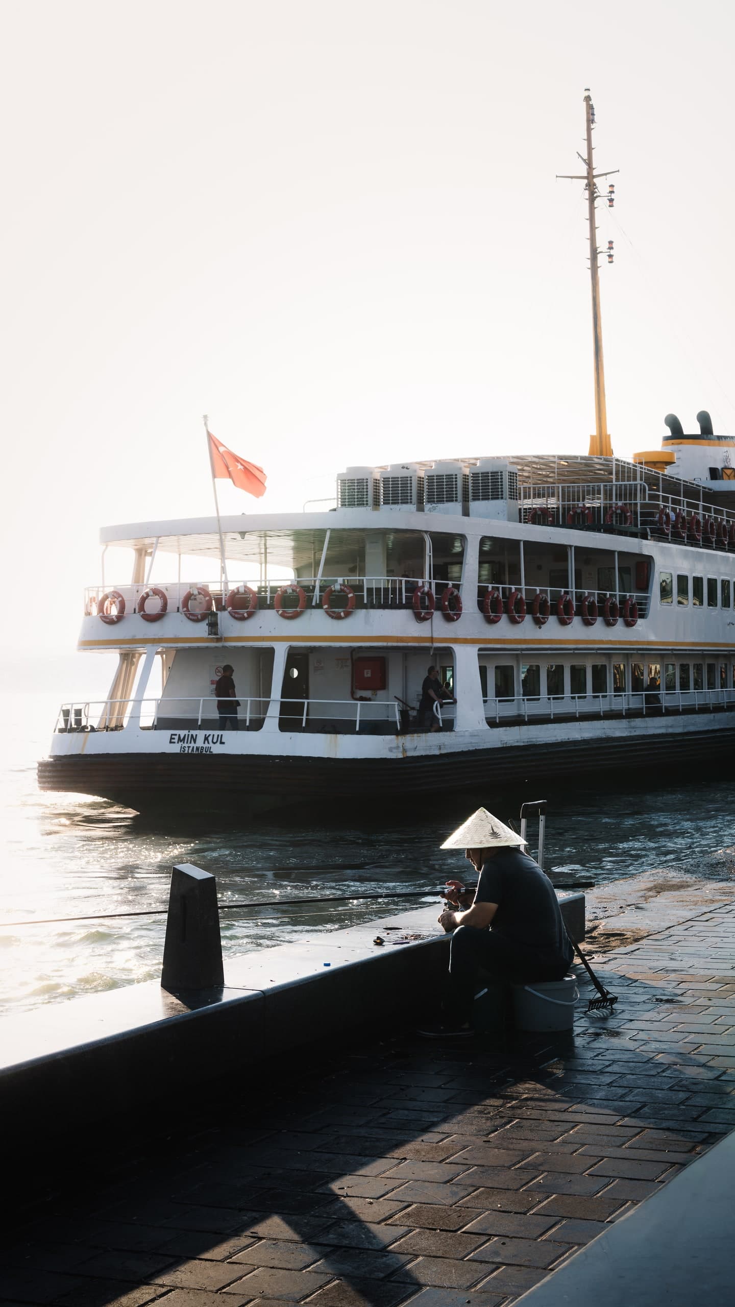 A ferry arriving in Istanbul as a fisherman sits on the dock in silhouette.