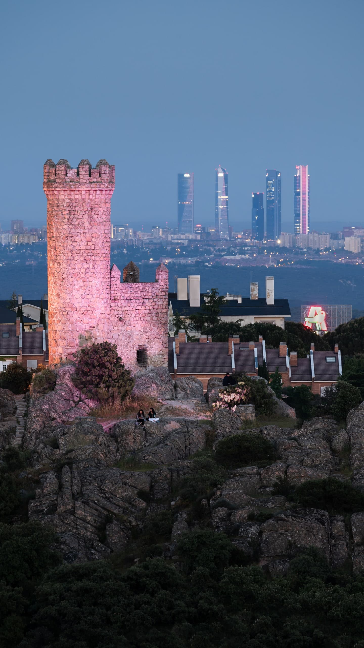 A ruined stone tower on a hillside, the Madrid skyline lit behind it at dusk.