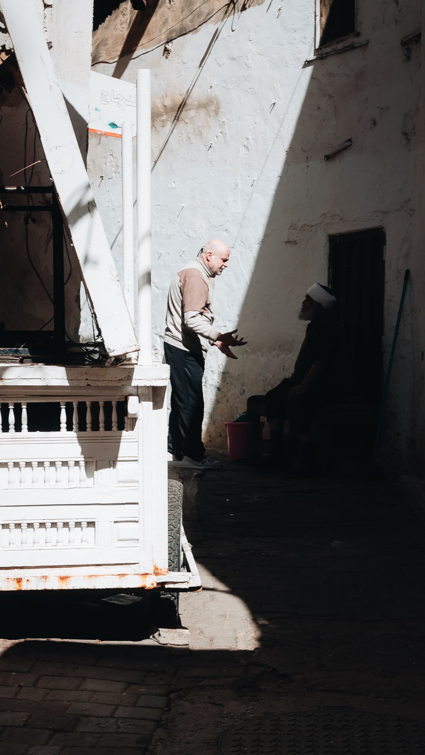 Two men talking in a sunlit Moroccan alley, sharp shadows cast against a white wall.