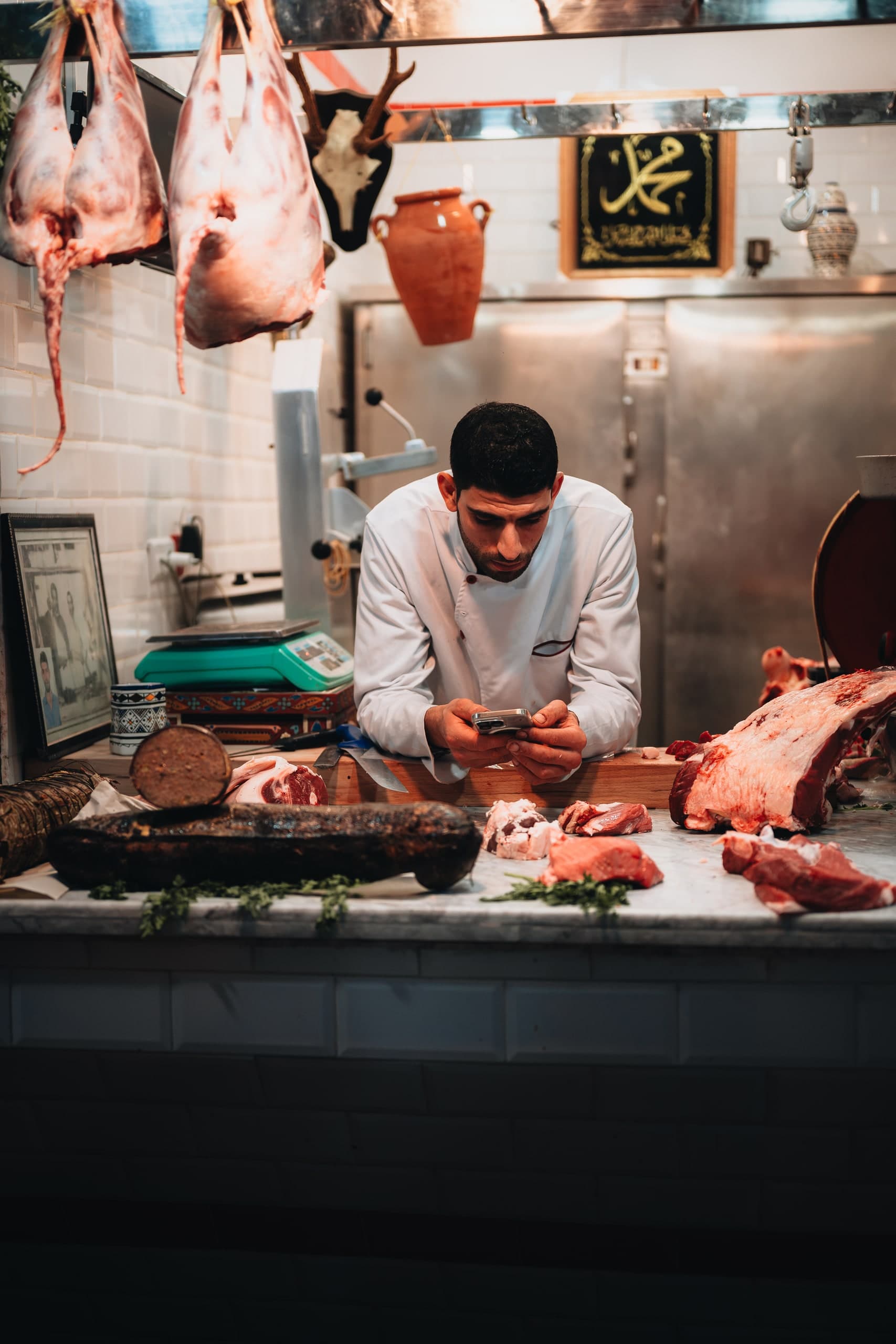 A butcher checking his phone at the counter, cuts of meat and a hanging carcass around him.