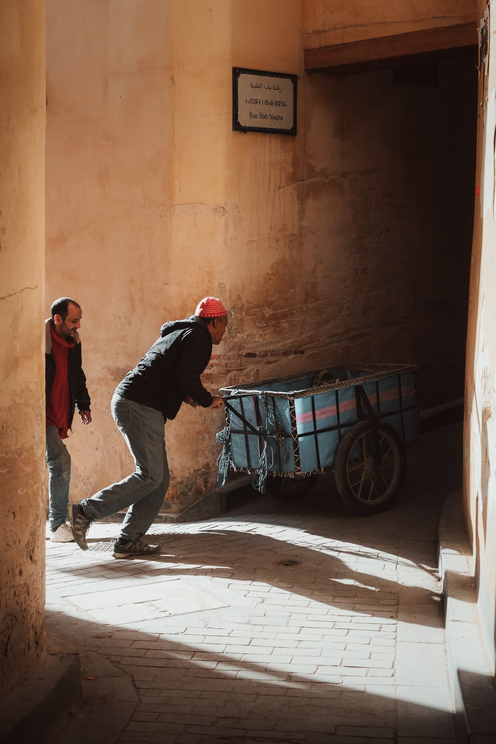 Two men pushing a loaded handcart through a sunlit passageway in a Moroccan medina.