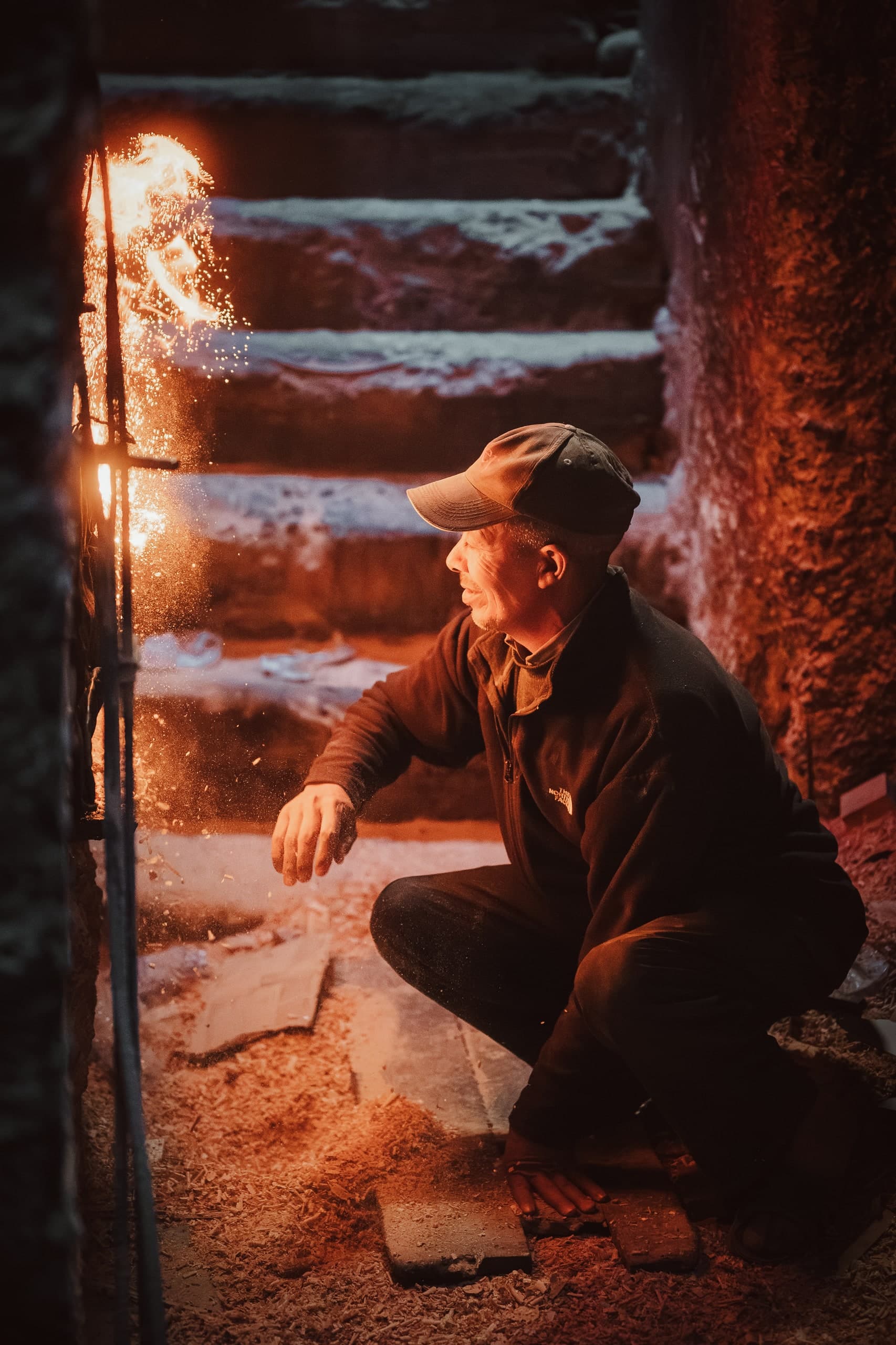 A man crouched beside a glowing fire on stone steps in a Moroccan workshop.