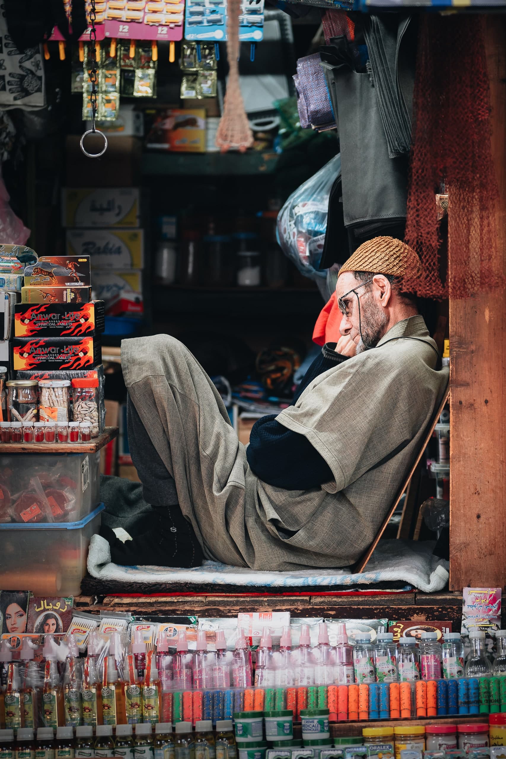 A shopkeeper reclining with his knees drawn up, resting in his stall surrounded by goods.