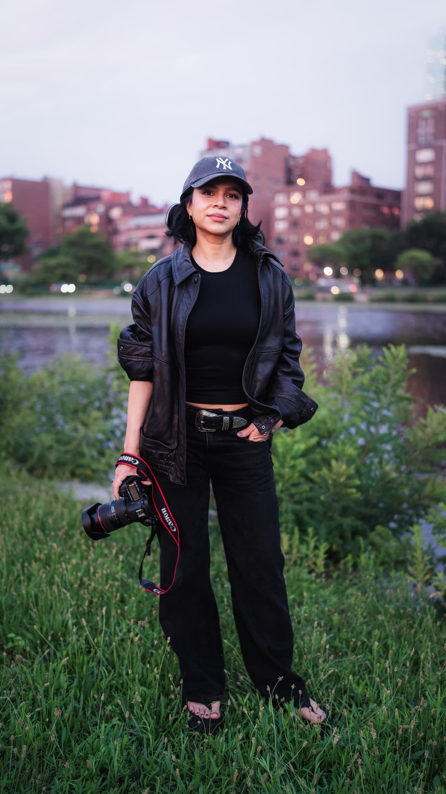 A photographer standing along the river at dusk, camera in hand, city skyline behind her.