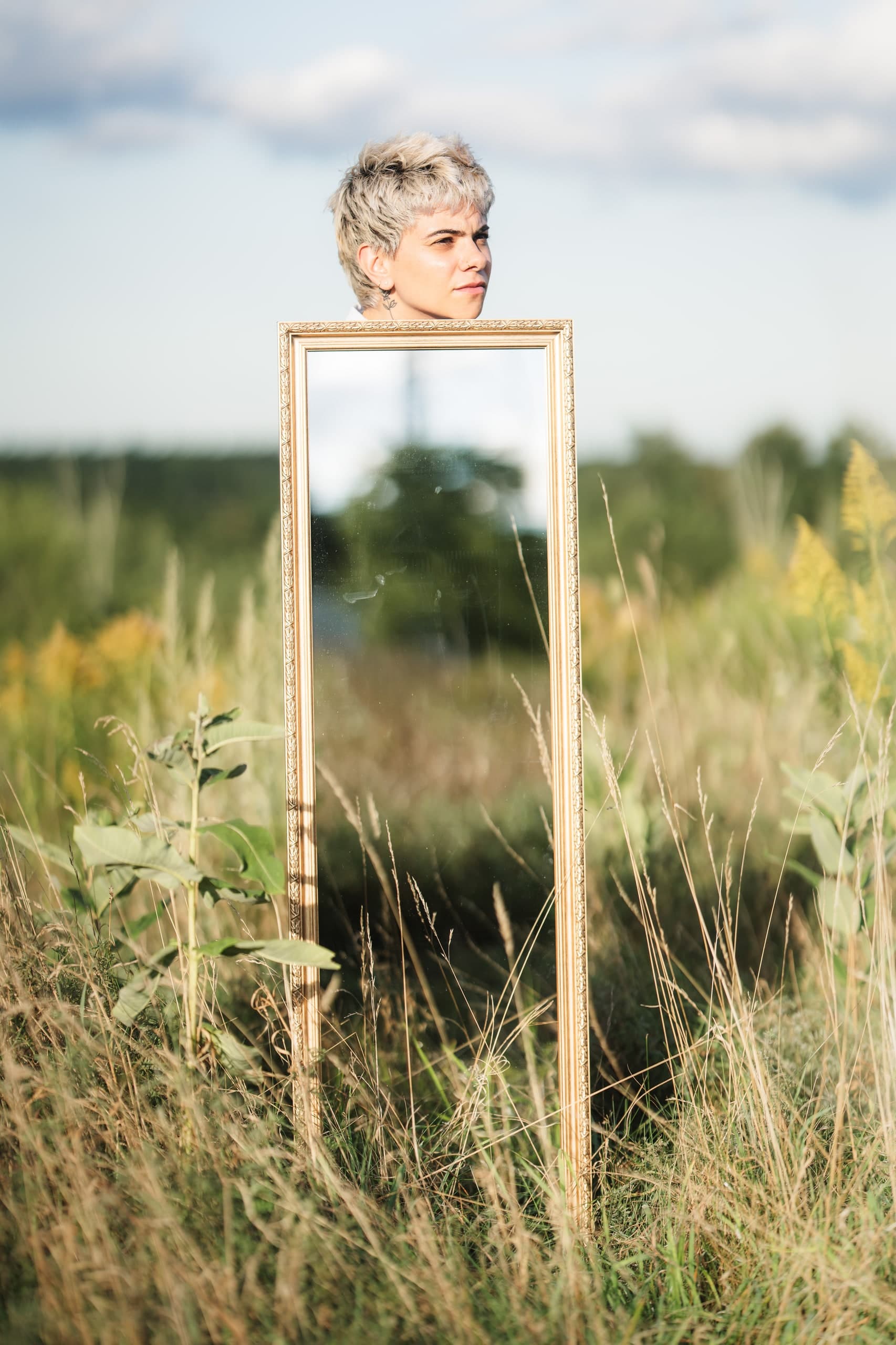 A woman's head visible above a freestanding mirror in a tall grass field, her reflection in the glass below.