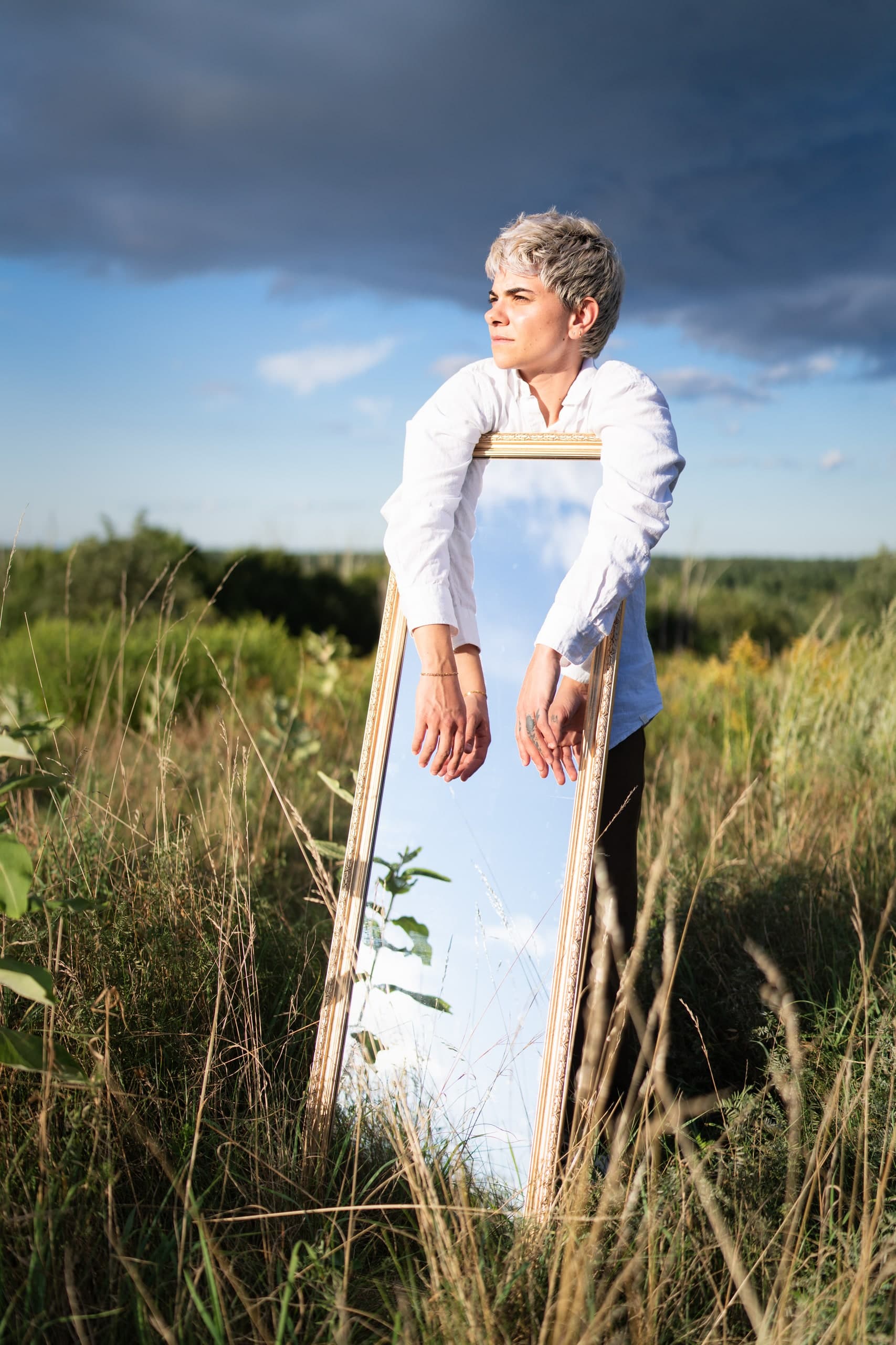 A woman leaning on a freestanding mirror in a tall grass field, storm clouds gathering behind her.