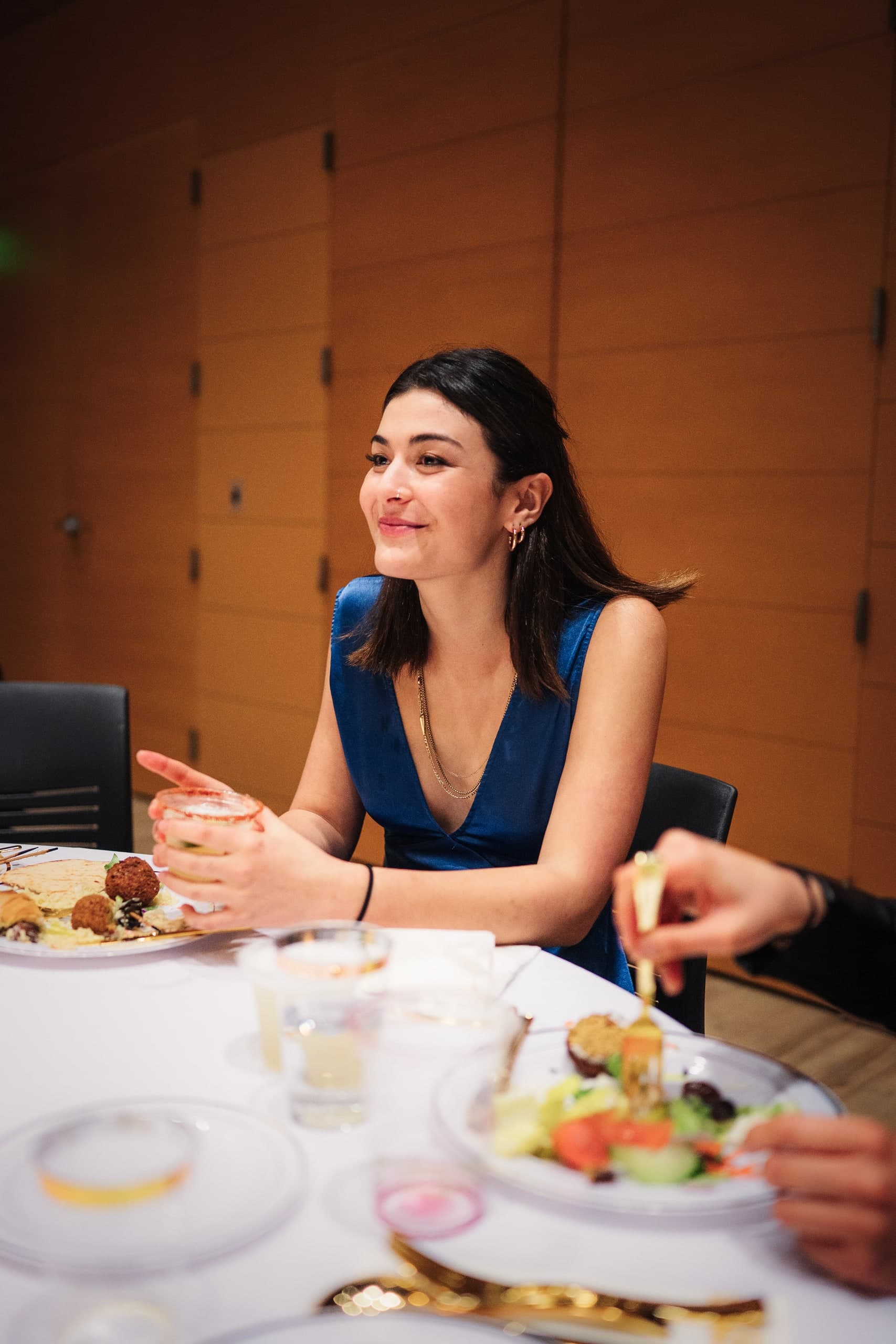 A woman sitting at a dining table, smiling and holding a glass, a salad beside her.