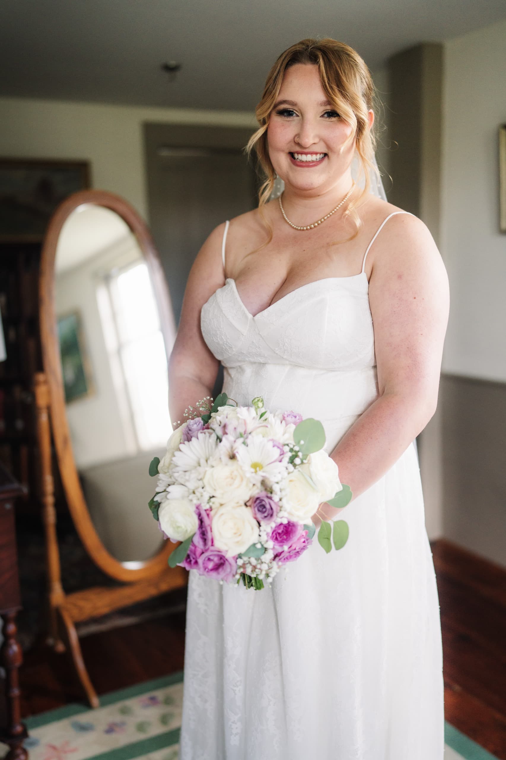 A bride smiling in front of a standing mirror, holding her bouquet of white and lavender flowers.