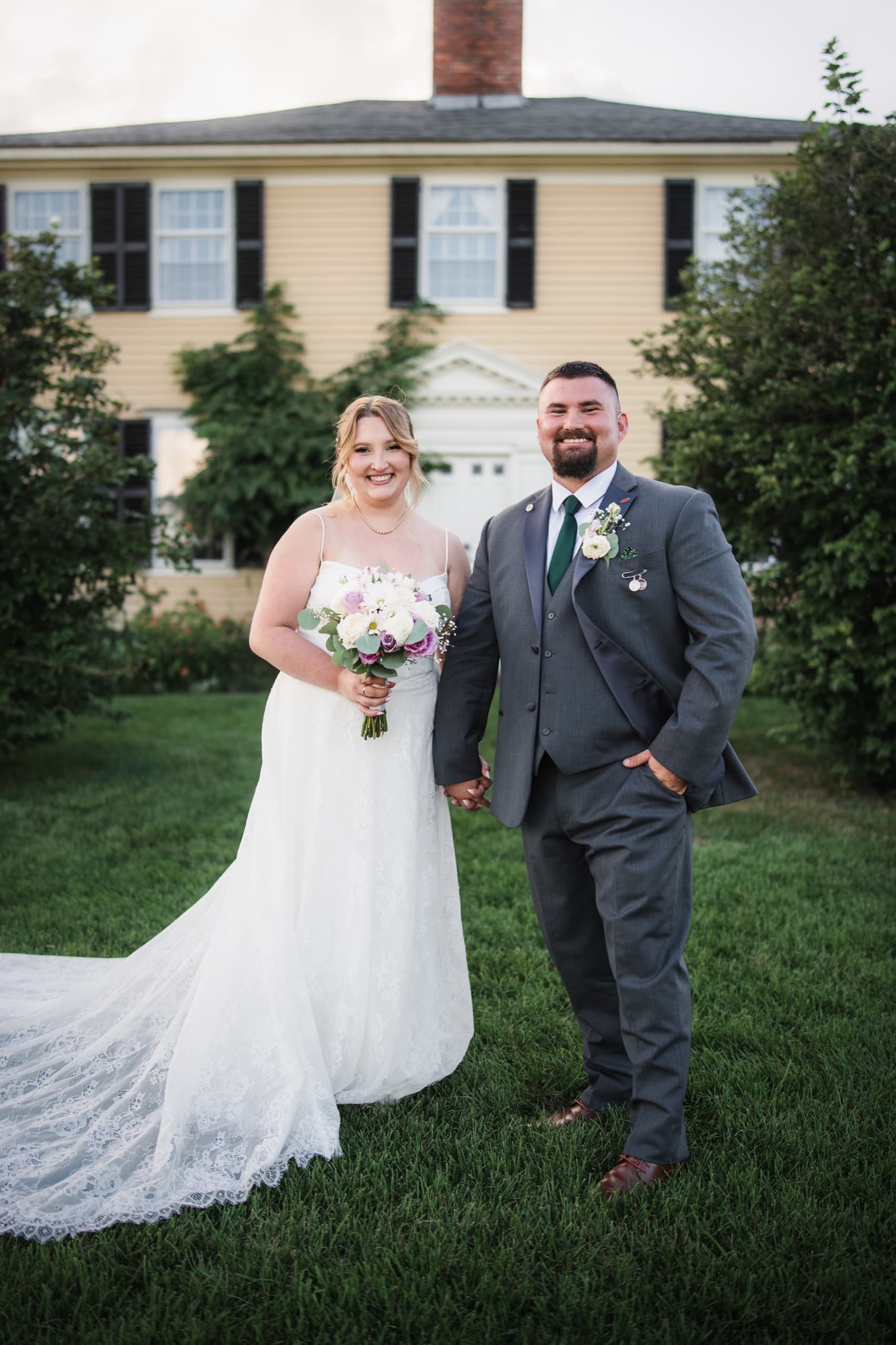 A newly married couple holding hands on the lawn in front of a colonial farmhouse.