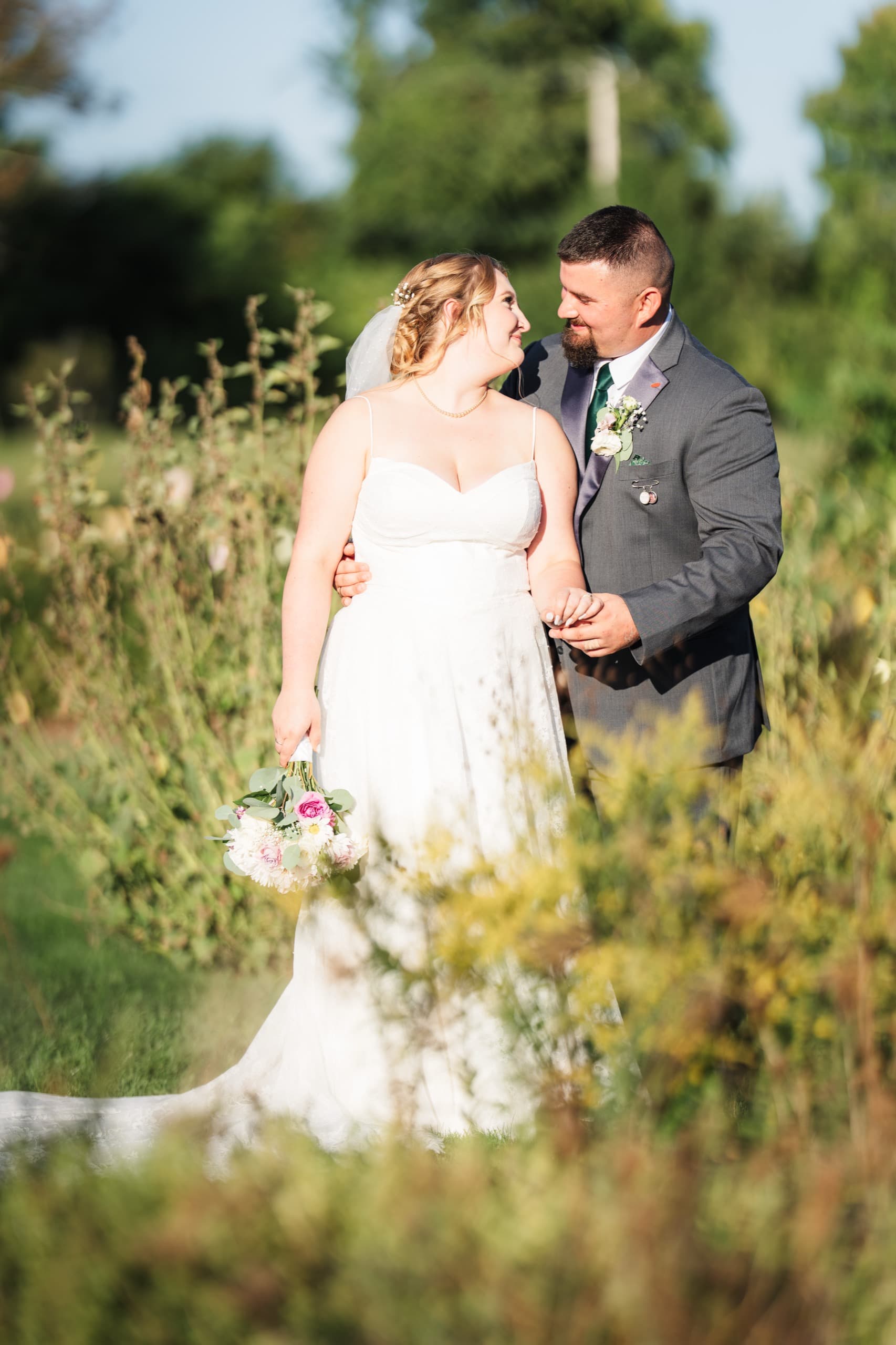 A bride and groom close together in a golden wildflower field, foreground grasses softly blurred.