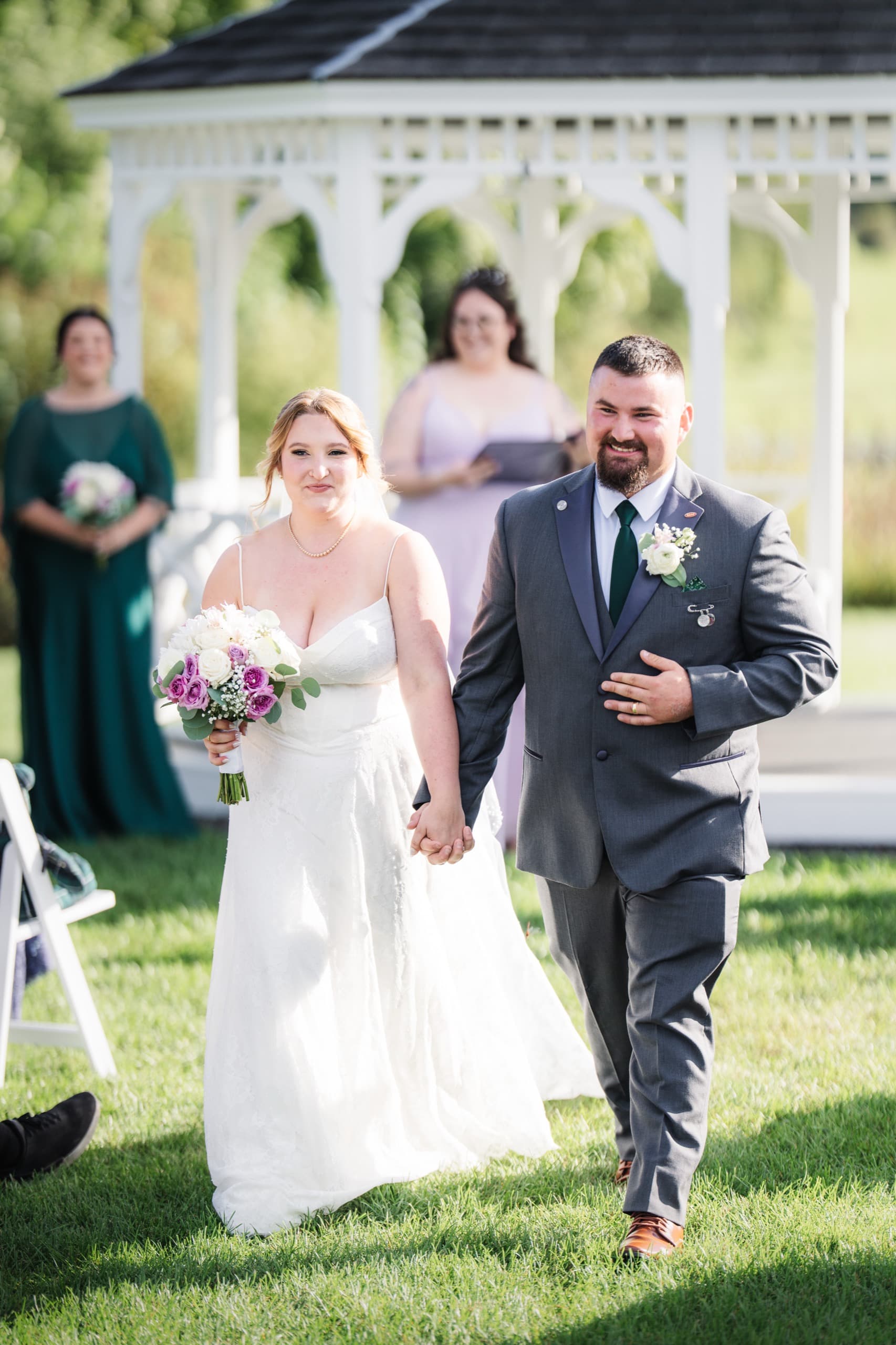 A newly married couple walking hand in hand past a white gazebo, both laughing.