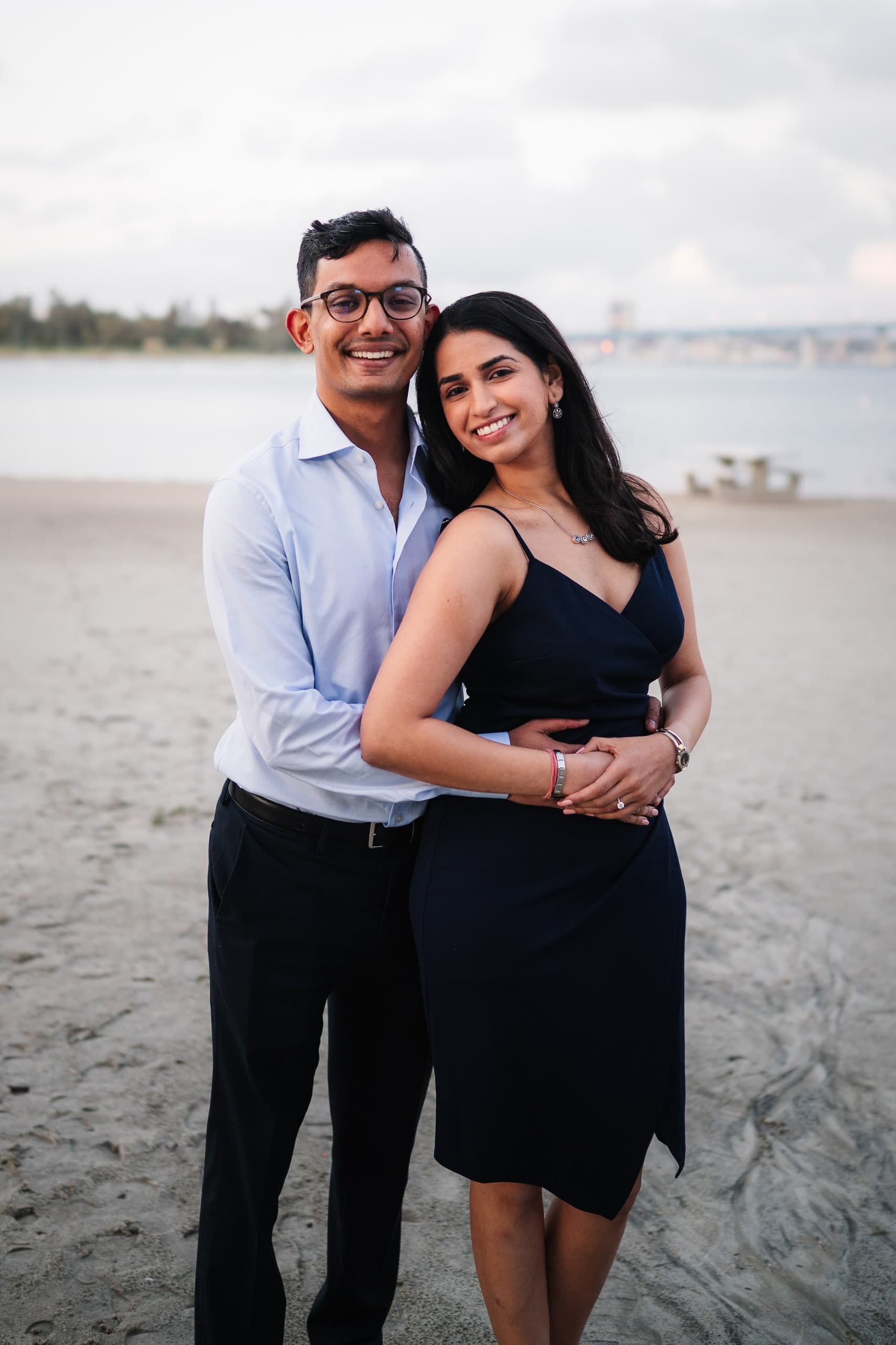 A couple embracing on a cloudy beach, a pier visible in the distance.