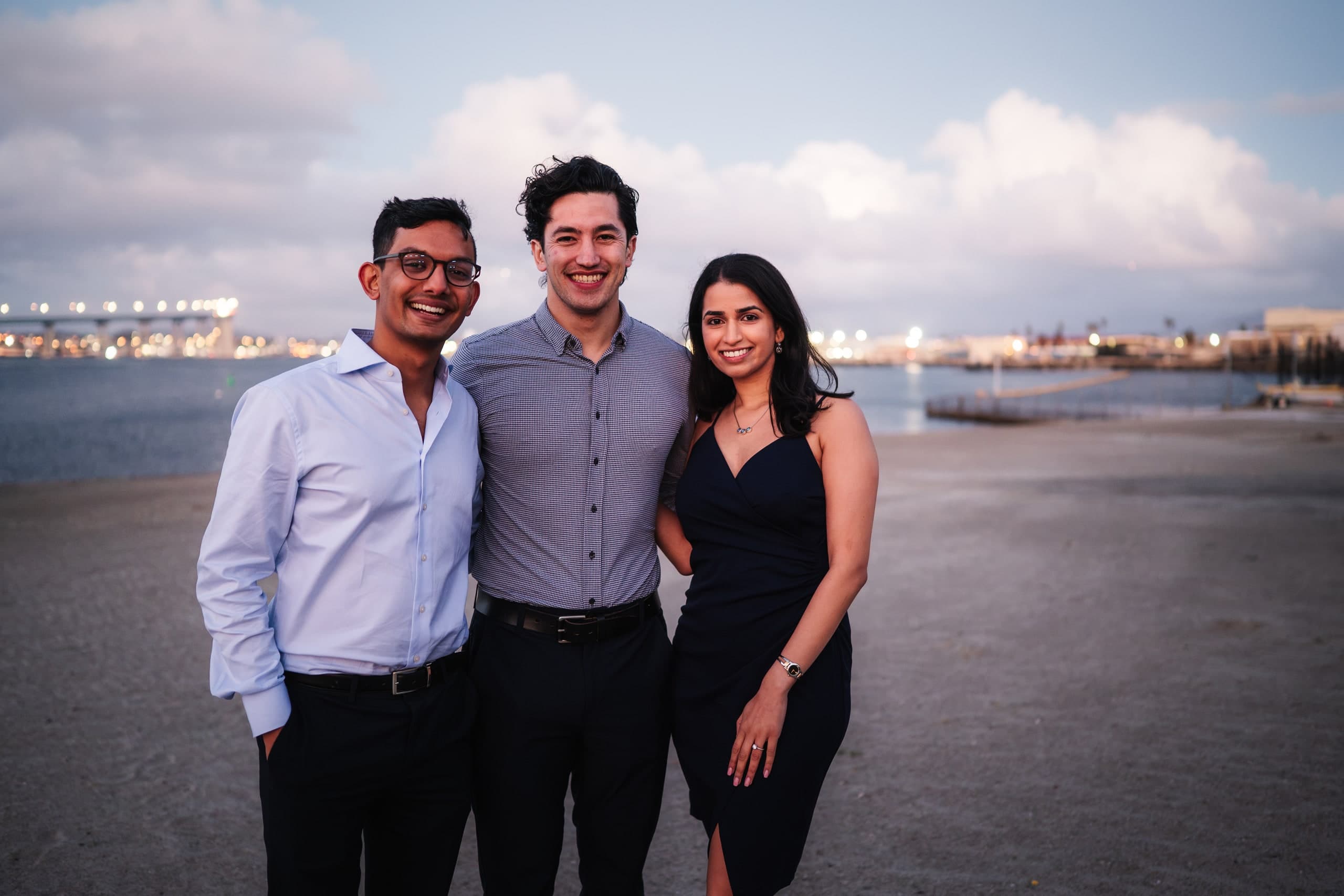 Three friends standing together on the beach at dusk, city lights across the water behind them.