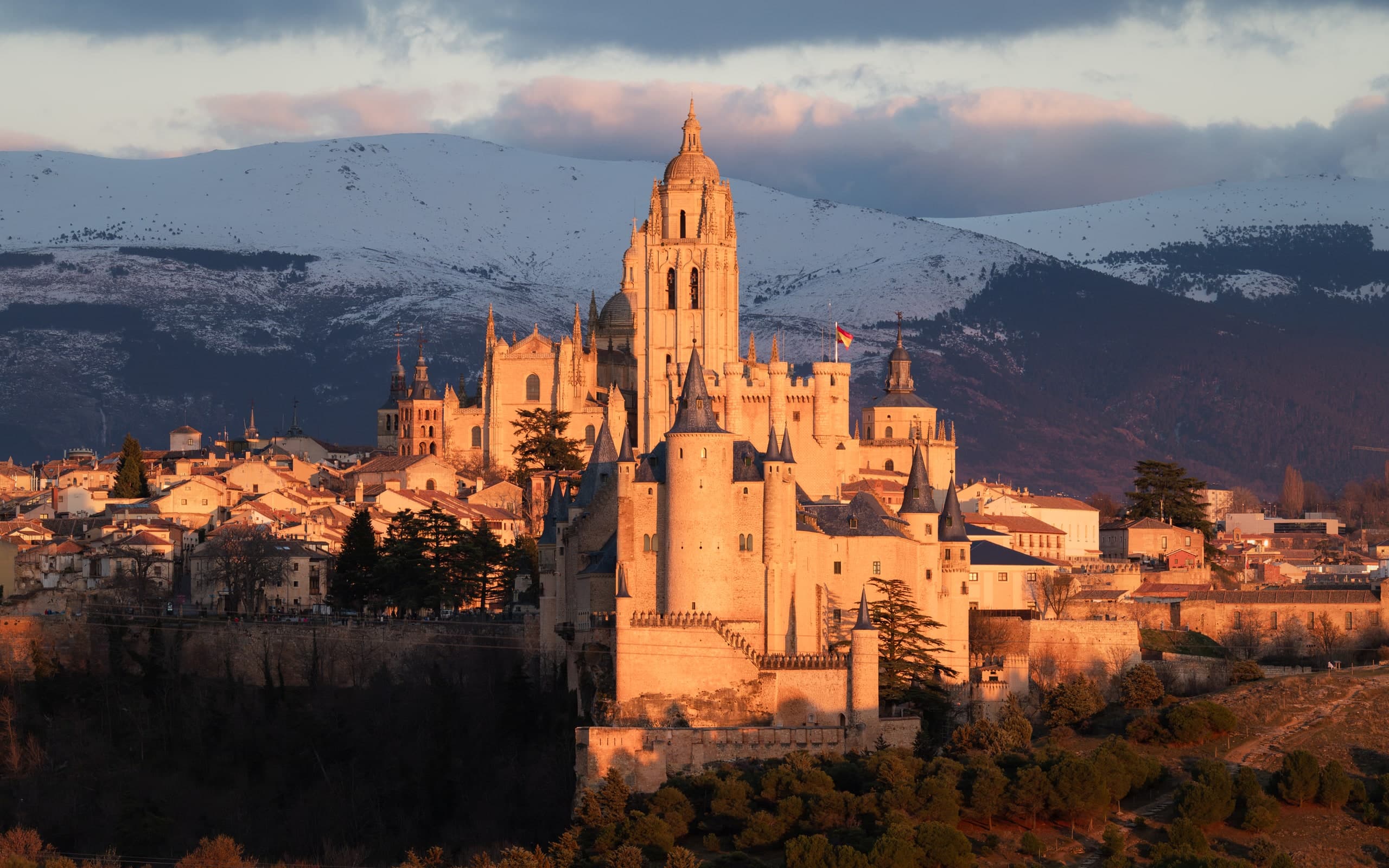 The Alcázar of Segovia at golden hour, snow-capped mountains rising behind the castle.