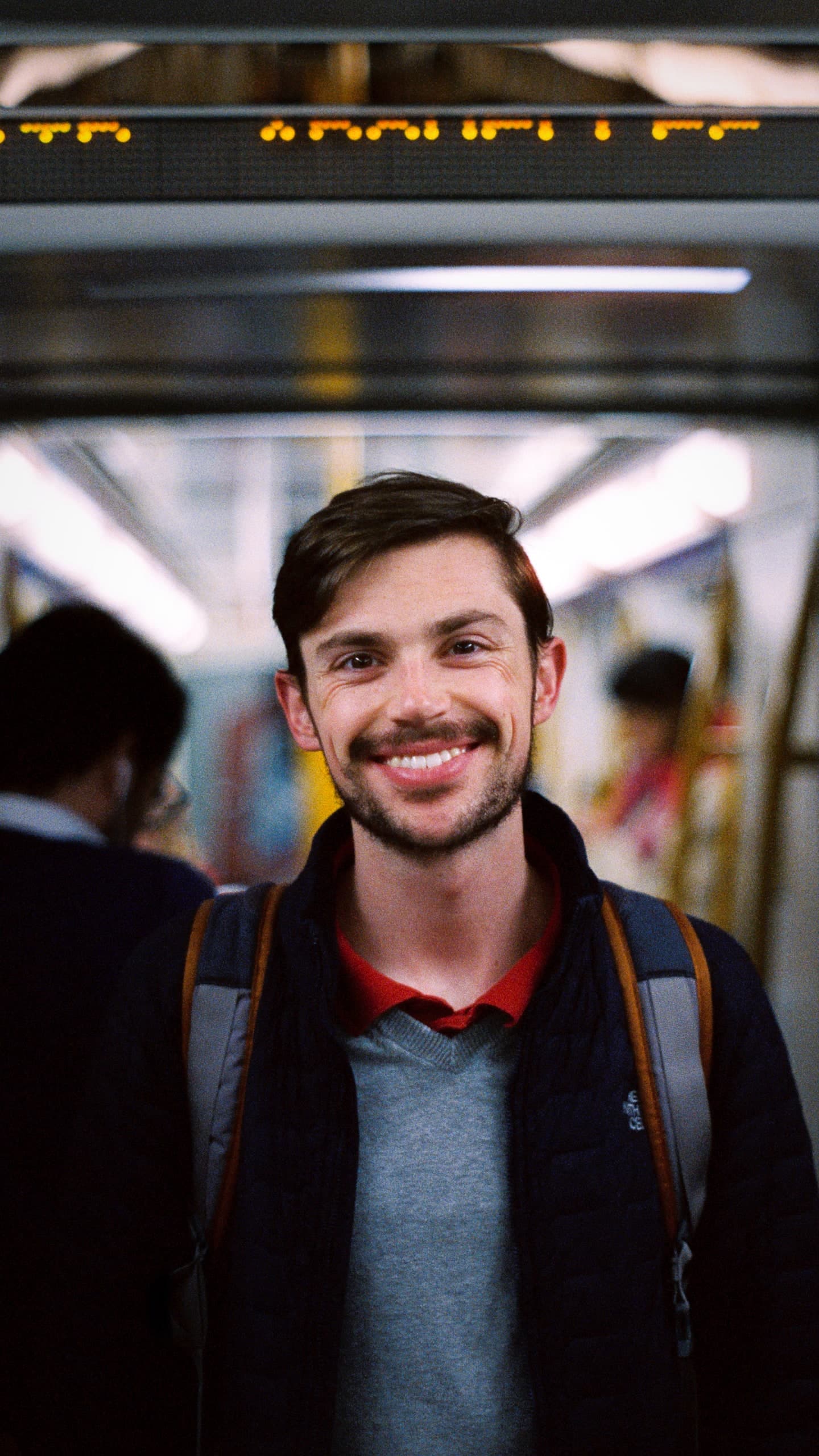 A young man smiling on a subway car, backpack straps on his shoulders, an arrivals board lit above him.