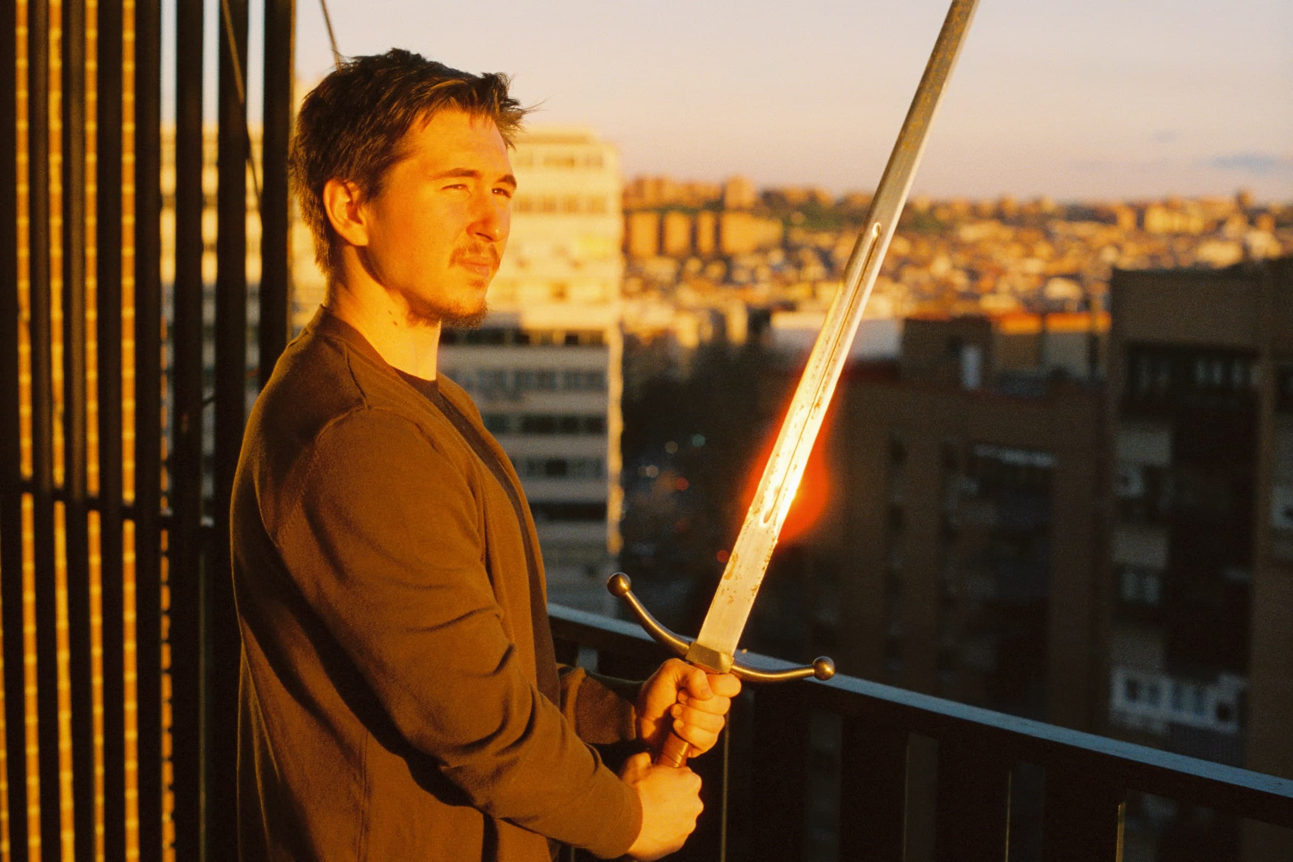 A man holding a sword up on a balcony at golden hour, light catching the blade, a city skyline behind him.