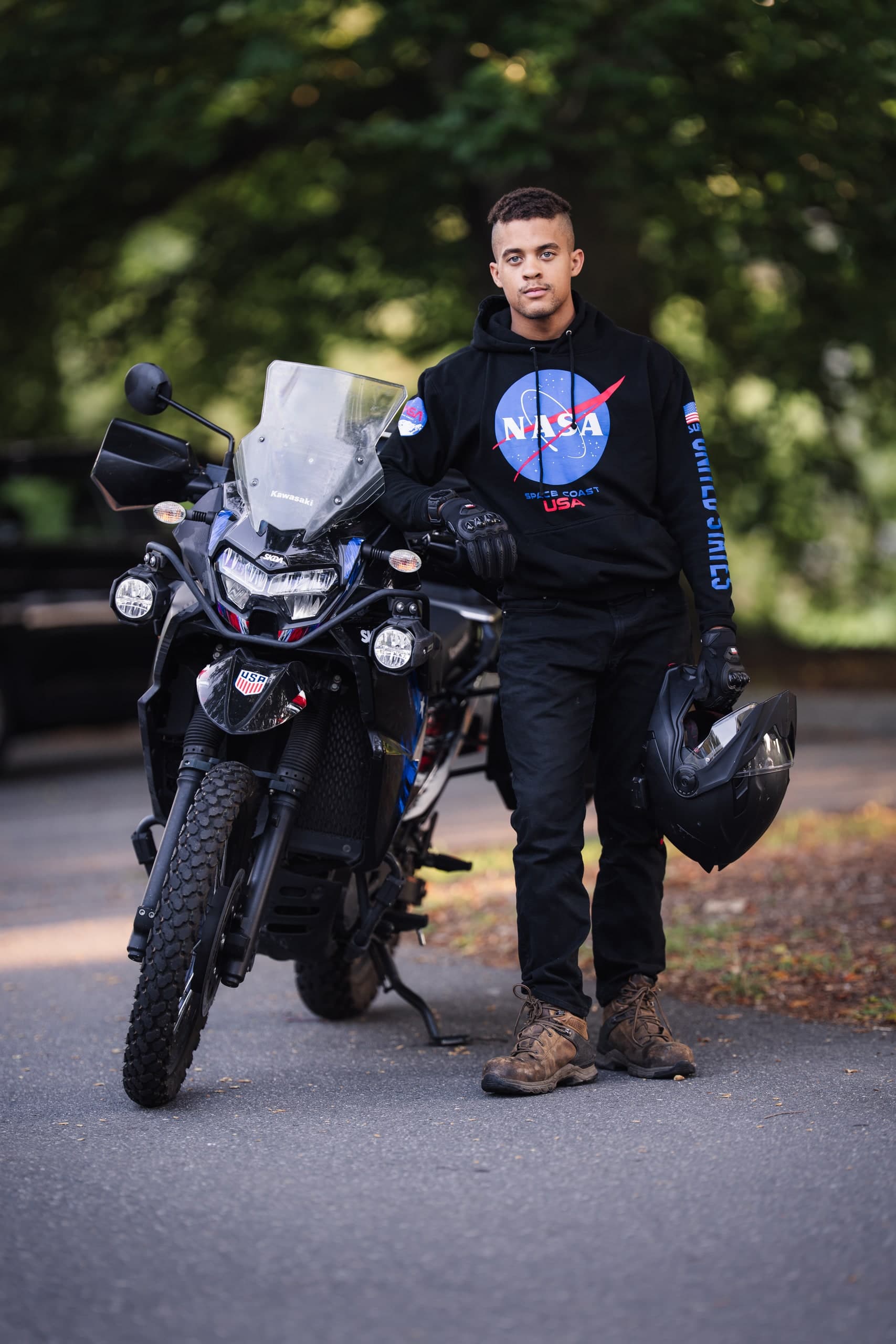 A young man in a NASA hoodie standing beside a motorcycle on a tree-lined road.