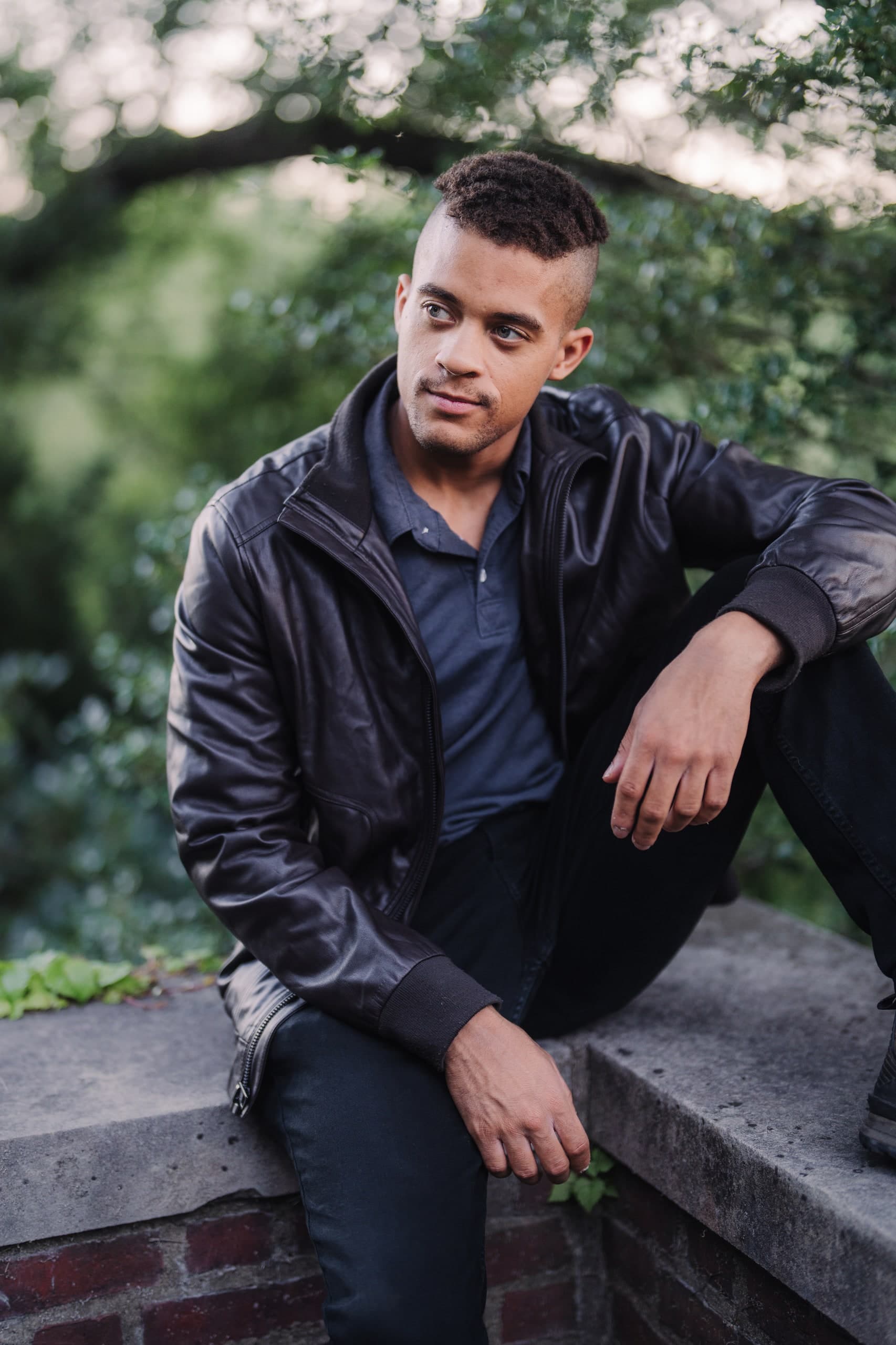 The same young man seated on a low garden wall, one leg drawn up, looking into the distance.